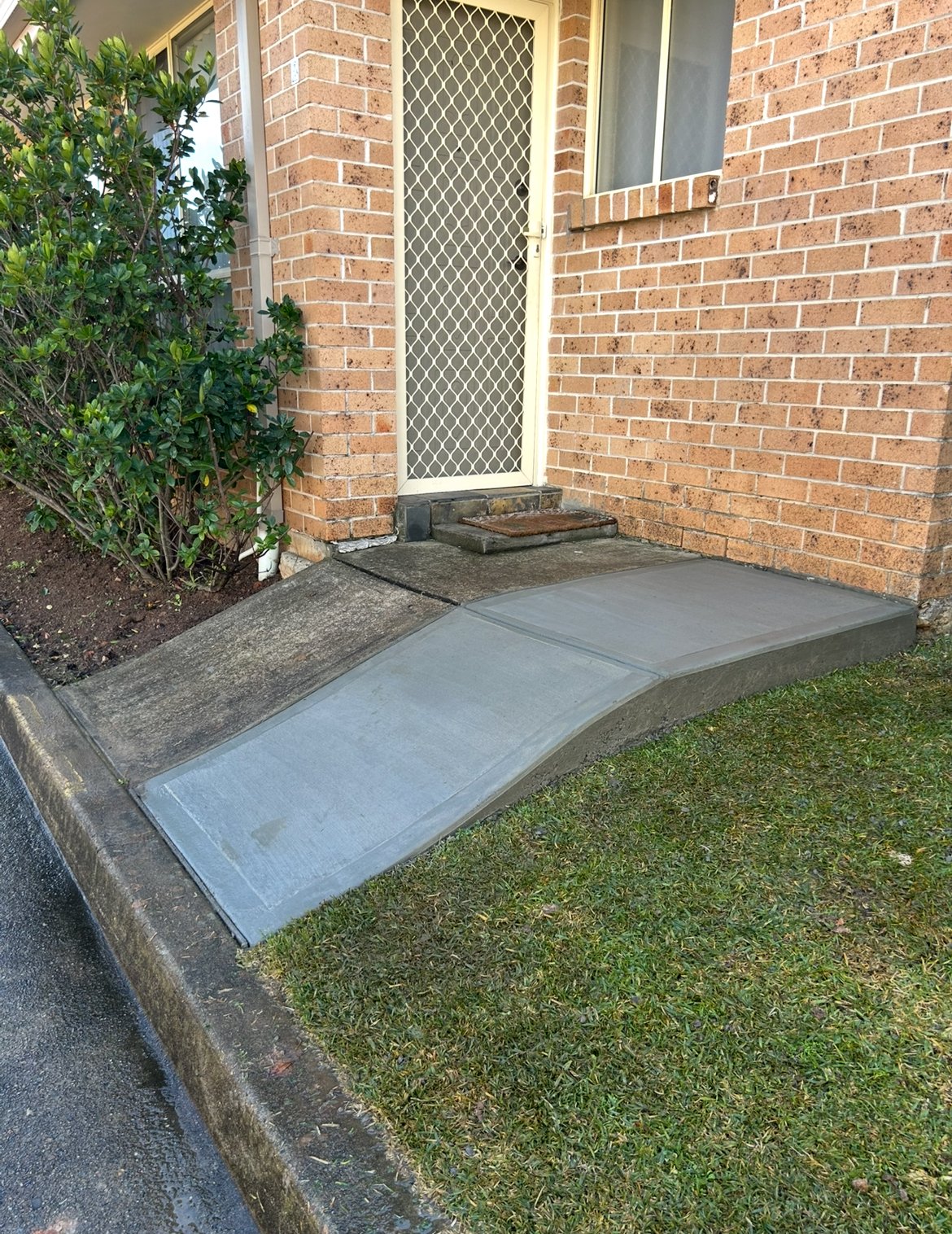 Concrete wheelchair ramp leading to the door of a brick house, with a doormat and a mesh security door.