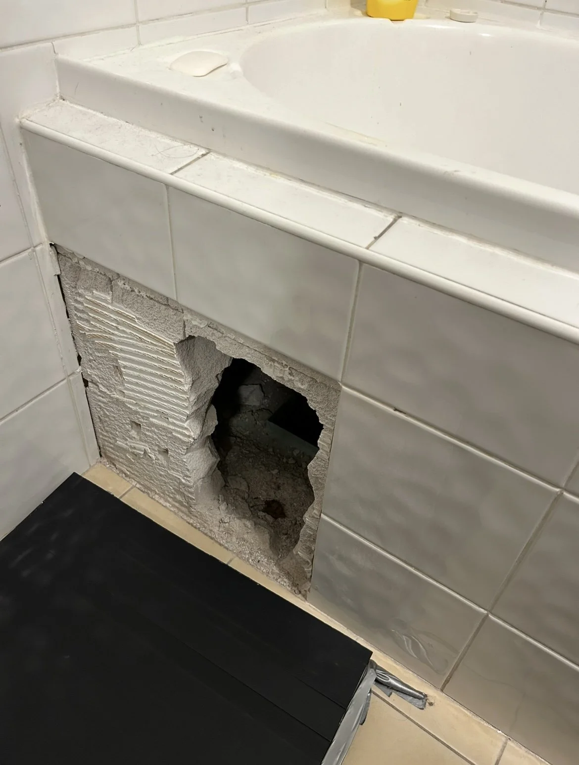 Bathroom wall with a section of damaged tile and exposed brick and drywall behind it near a bathtub.