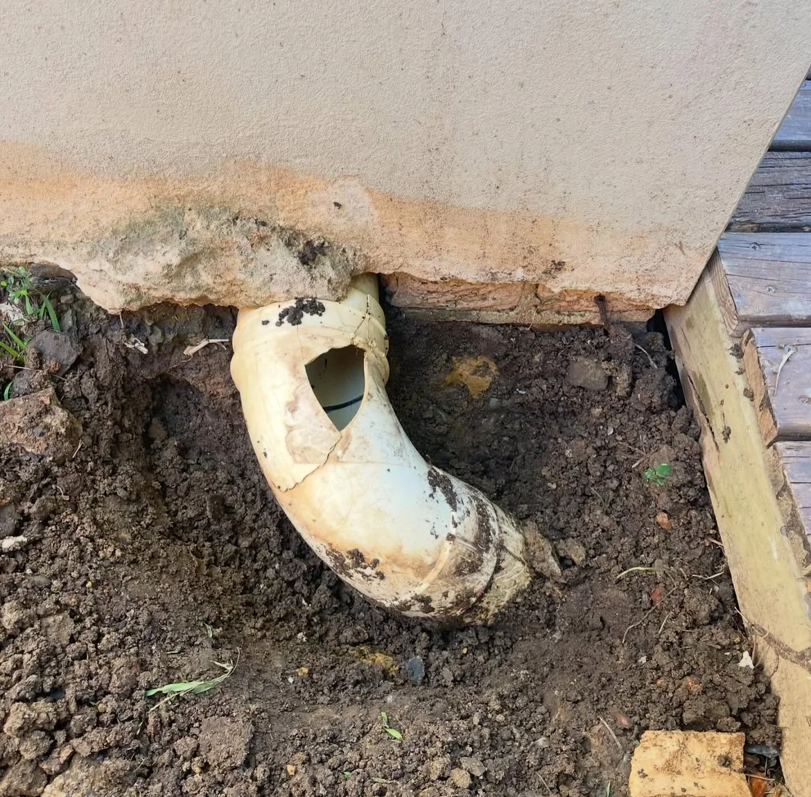 A broken ceramic pipe sticking out of the ground near a wall and a wooden deck.