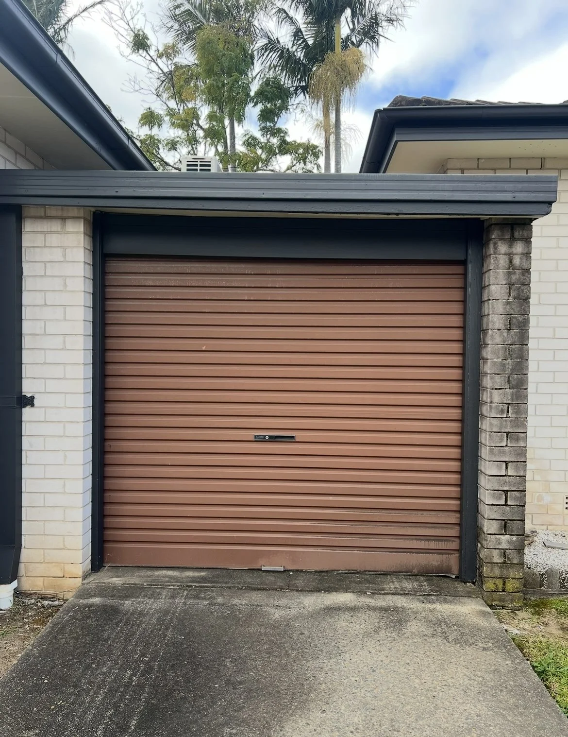 Closed brown roll-up garage door with concrete driveway in front and brick walls on each side, some trees and blue sky above.