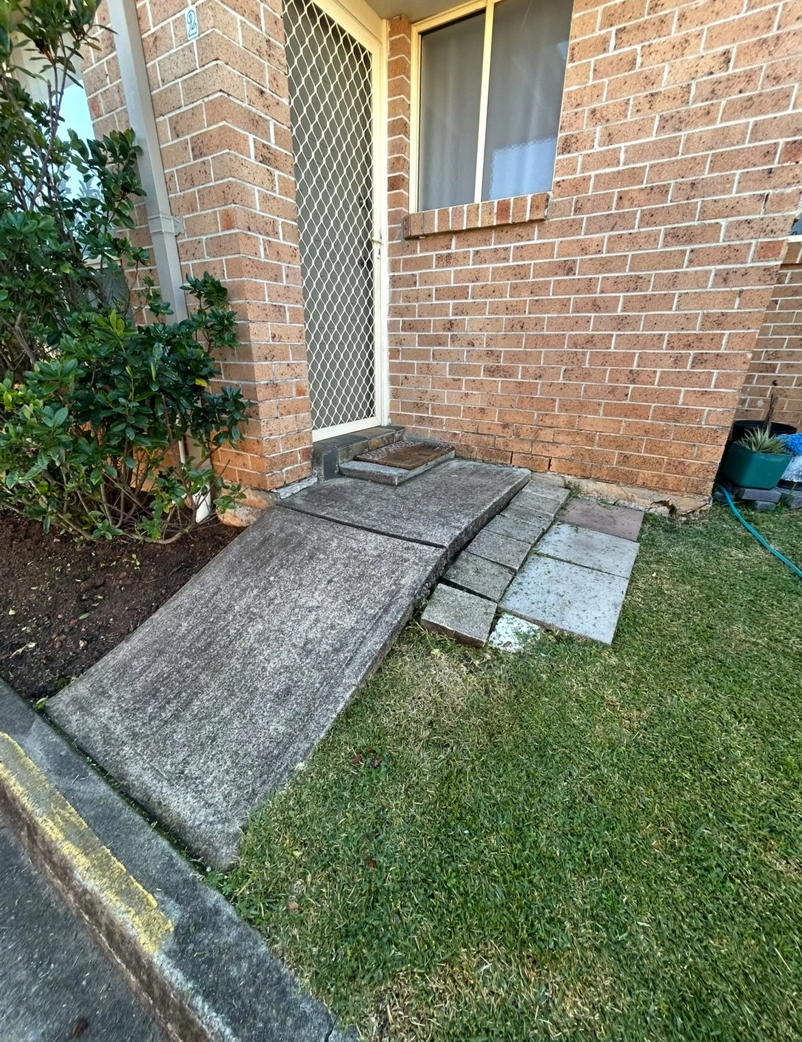 A small concrete ramp leading up to a door with a screen door on a brick house. There are step stones on the ground to the right of the ramp, and a small patch of grass in front of it. A bush is on the left side of the image, and a garden hose is on the right side near a gardening container.