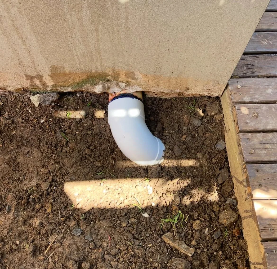 White PVC pipe elbow fitting installed below the exterior wall of a house, with dirt and small plants in the soil around it, adjacent to a wooden deck.