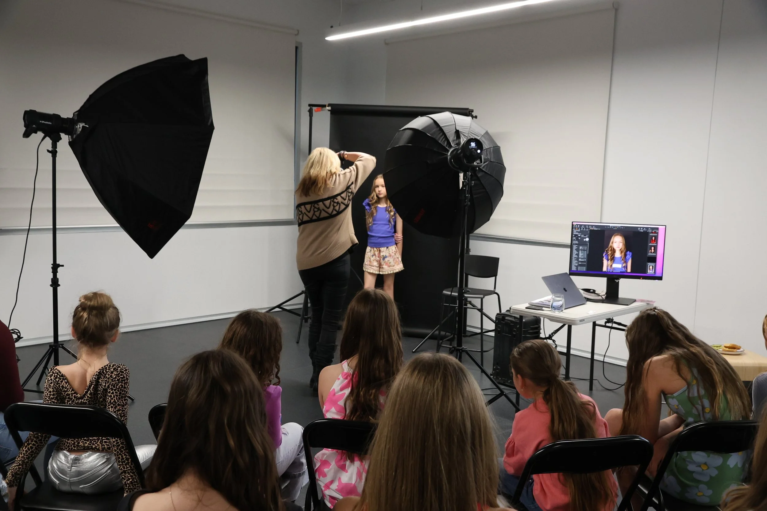 Photography studio with a photographer and model being photographed. Several children are seated, watching the session. Studio lighting, a black backdrop, and a computer monitor displaying the photo are visible.
