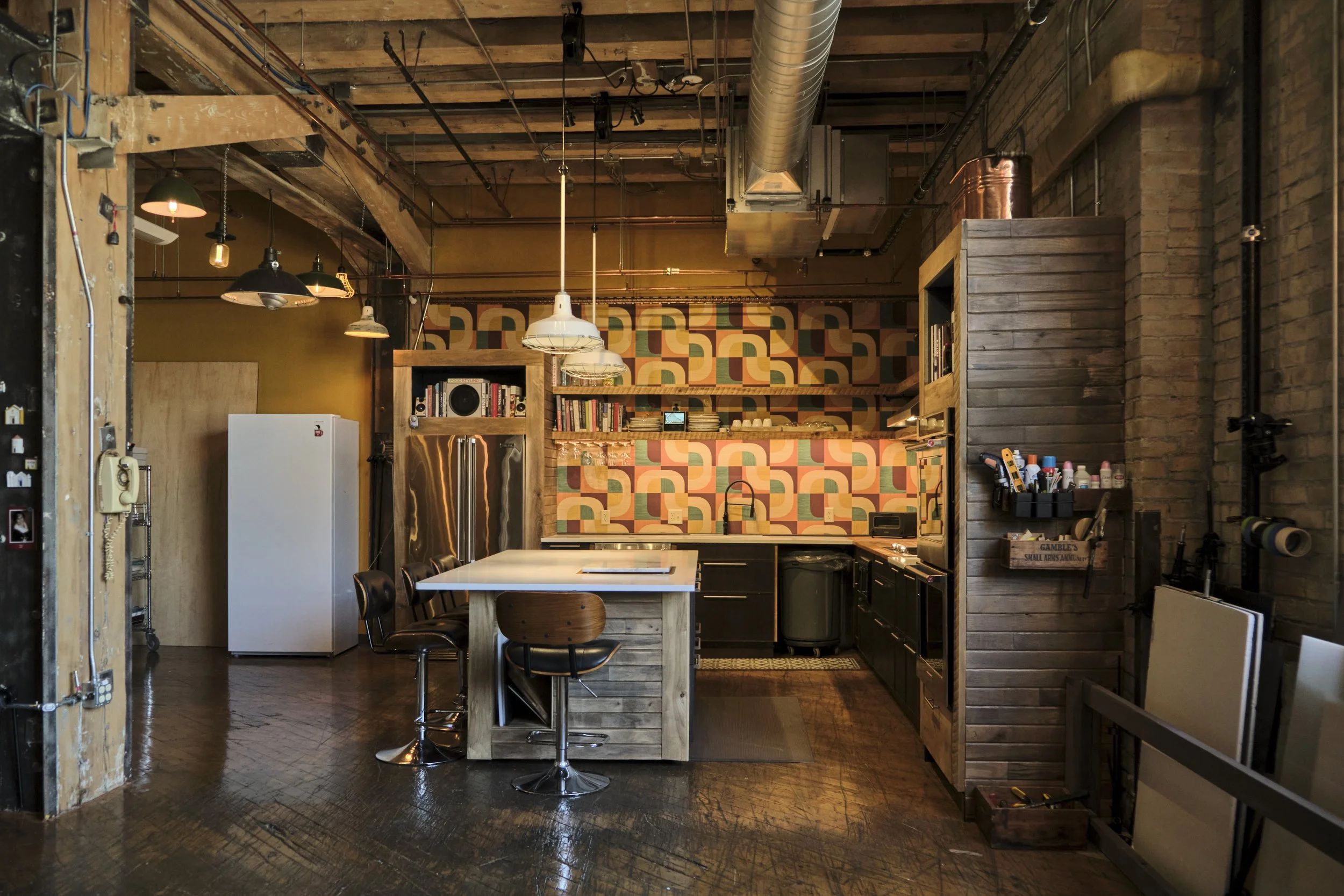 Modern kitchen with geometric patterned backsplash, black cabinetry, wooden accents, and hanging pendant lights in an industrial-style open space.