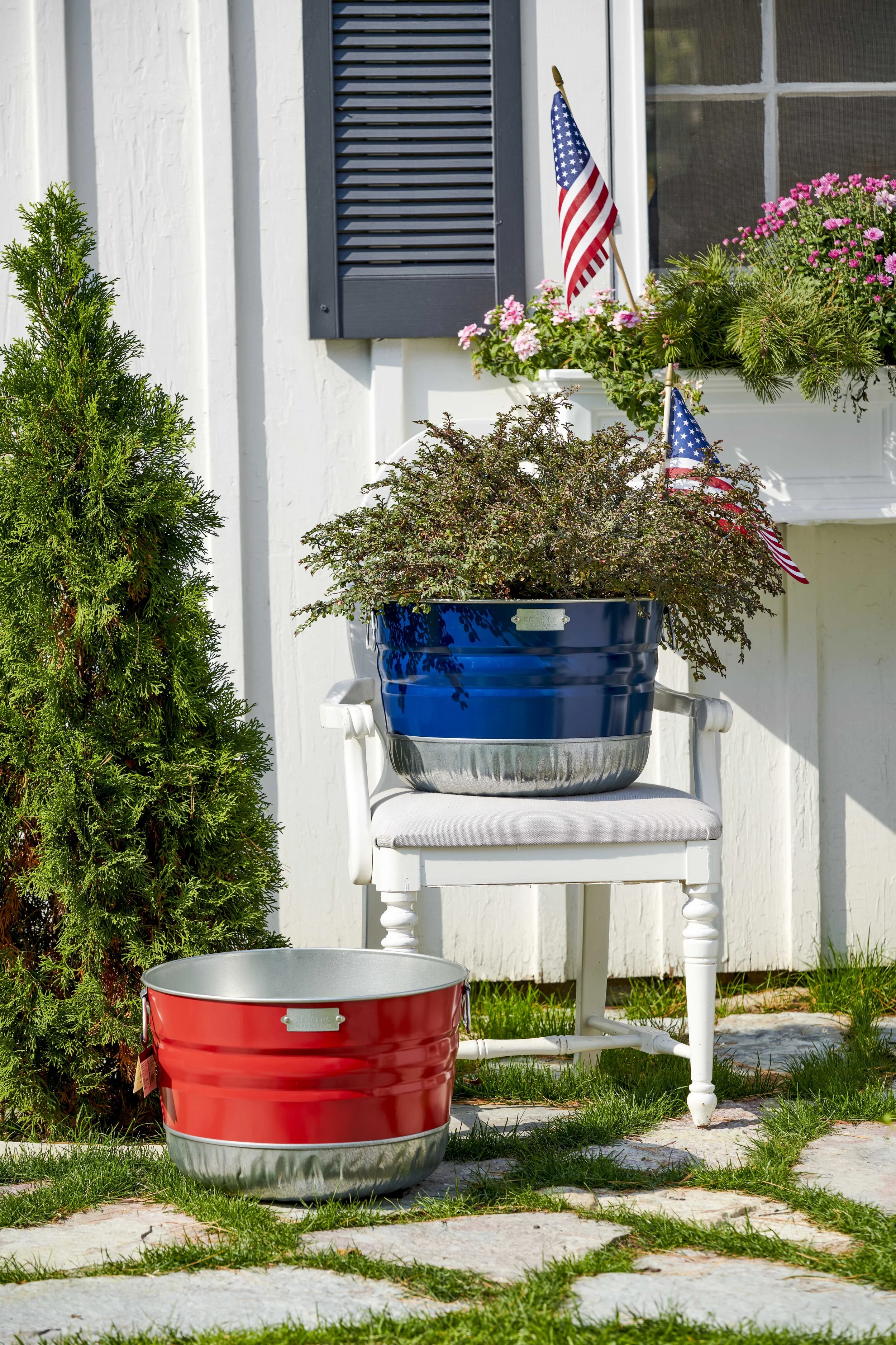 Decorative outdoor display with two patriotic-themed metal buckets, one red and one blue, on and beside a white wooden chair. The blue bucket on the chair contains a potted plant with small purple-green leaves. The background features a white house w