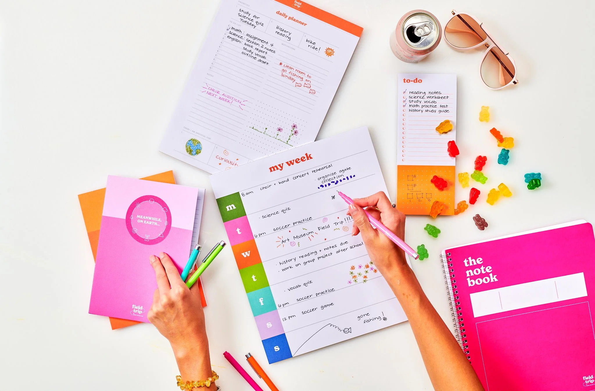 Images of school supplies and planning pages on a white desk. There are colorful notebooks, a weekly planner, a daily planner, a to-do list, pens, gummy bears, a soda can, and pink sunglasses.