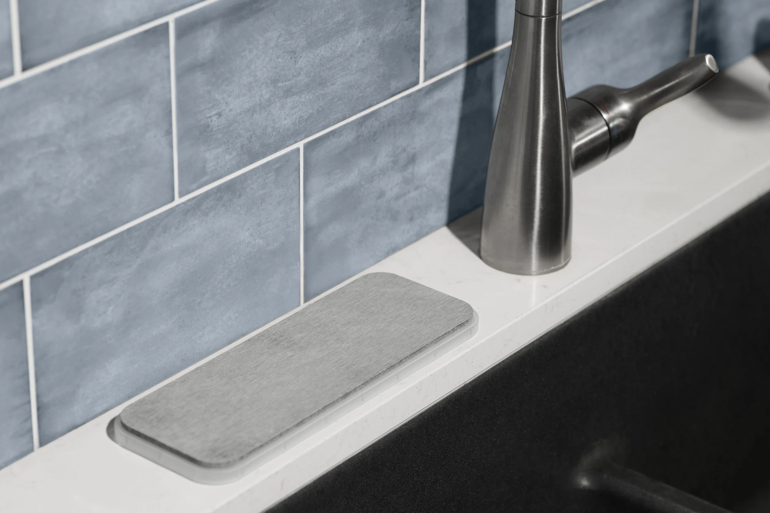 Close-up of a modern kitchen sink area with a stainless steel faucet, blue tiled backsplash, and a light gray cutting board on the white countertop.