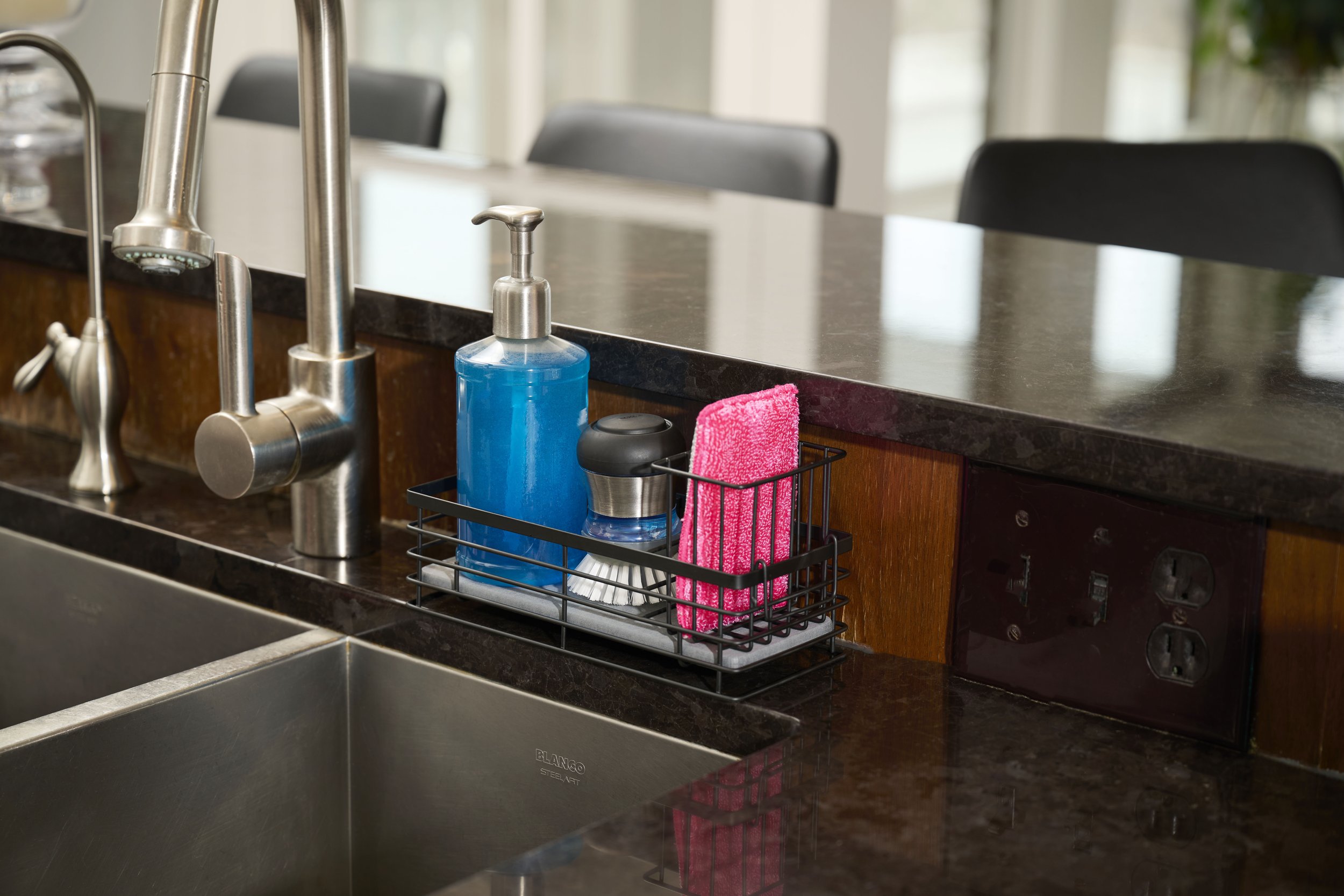 Kitchen sink with soap dispenser, hand sanitizer, and pink cloth in a wire basket on dark granite countertop.