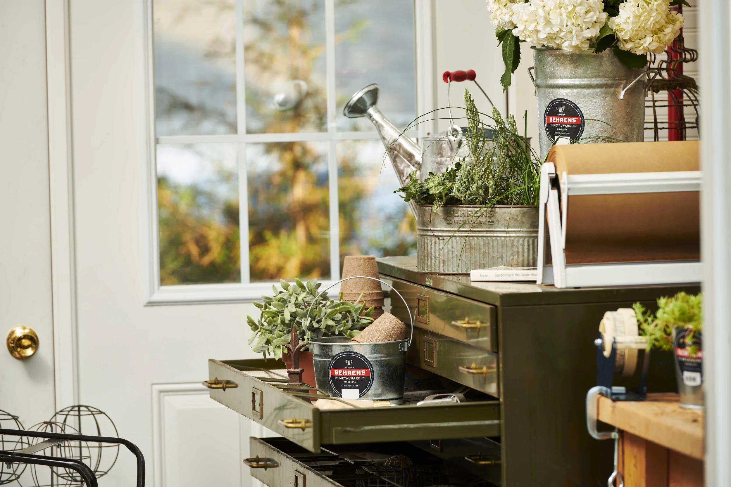 Indoor scene with gardening supplies on a vintage metal cabinet near a window, including potted plants, gardening tools, and plant markers.