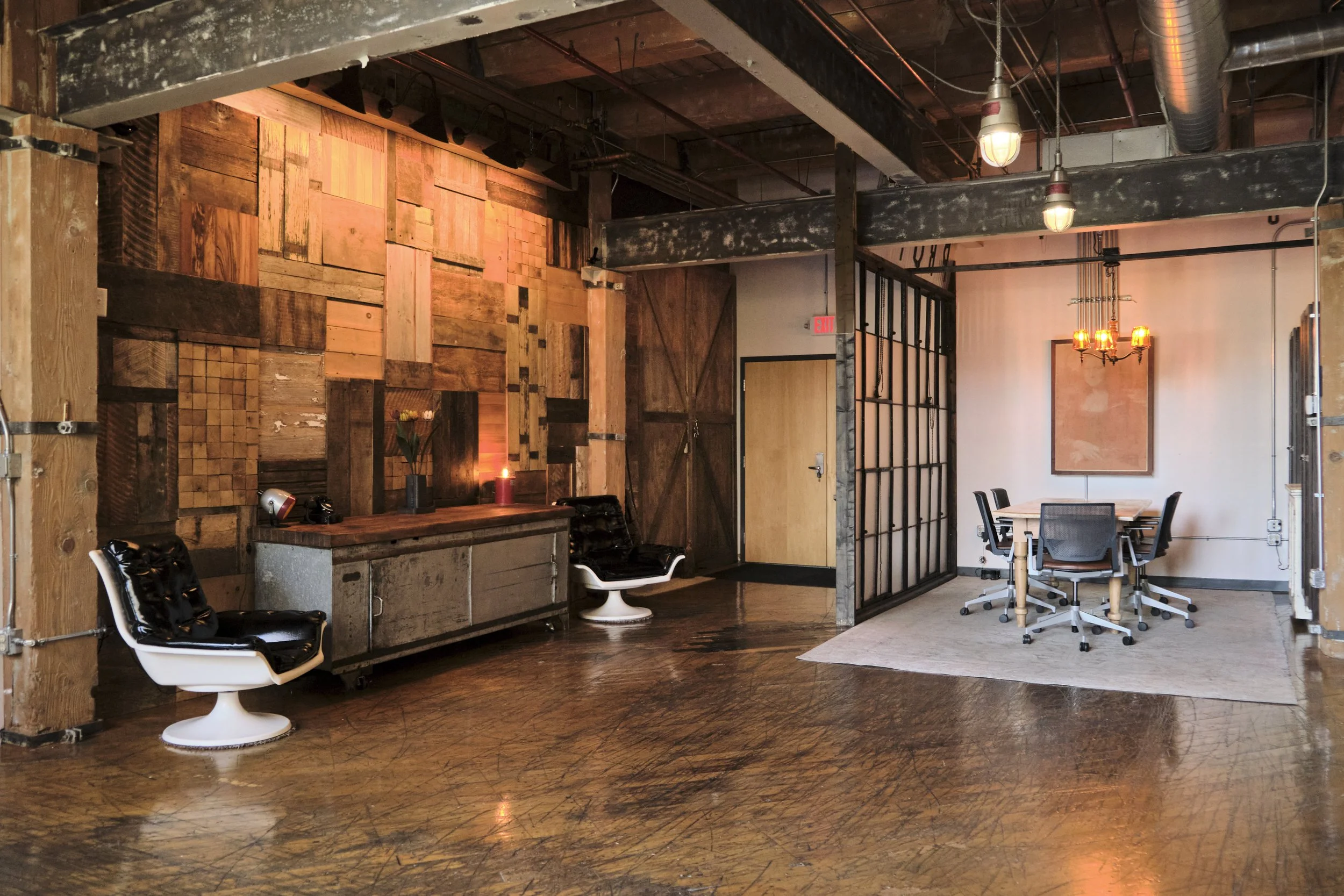 An industrial-style office space with a wooden accent wall, vintage chairs, a conference table, and exposed ductwork and lighting fixtures.