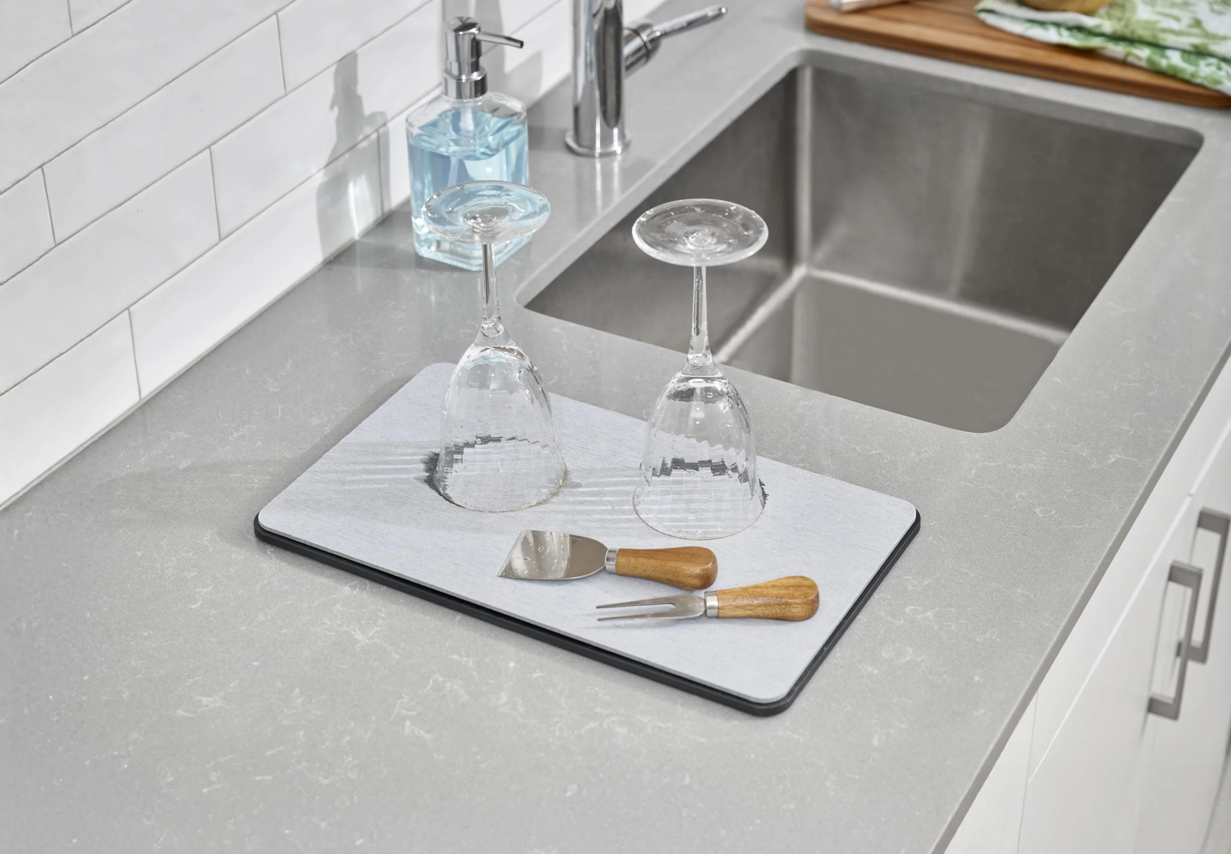 A kitchen countertop with a drying board holding two upside-down wine glasses, a bottle of blue liquid soap, and a ice scraper with a wooden handle.