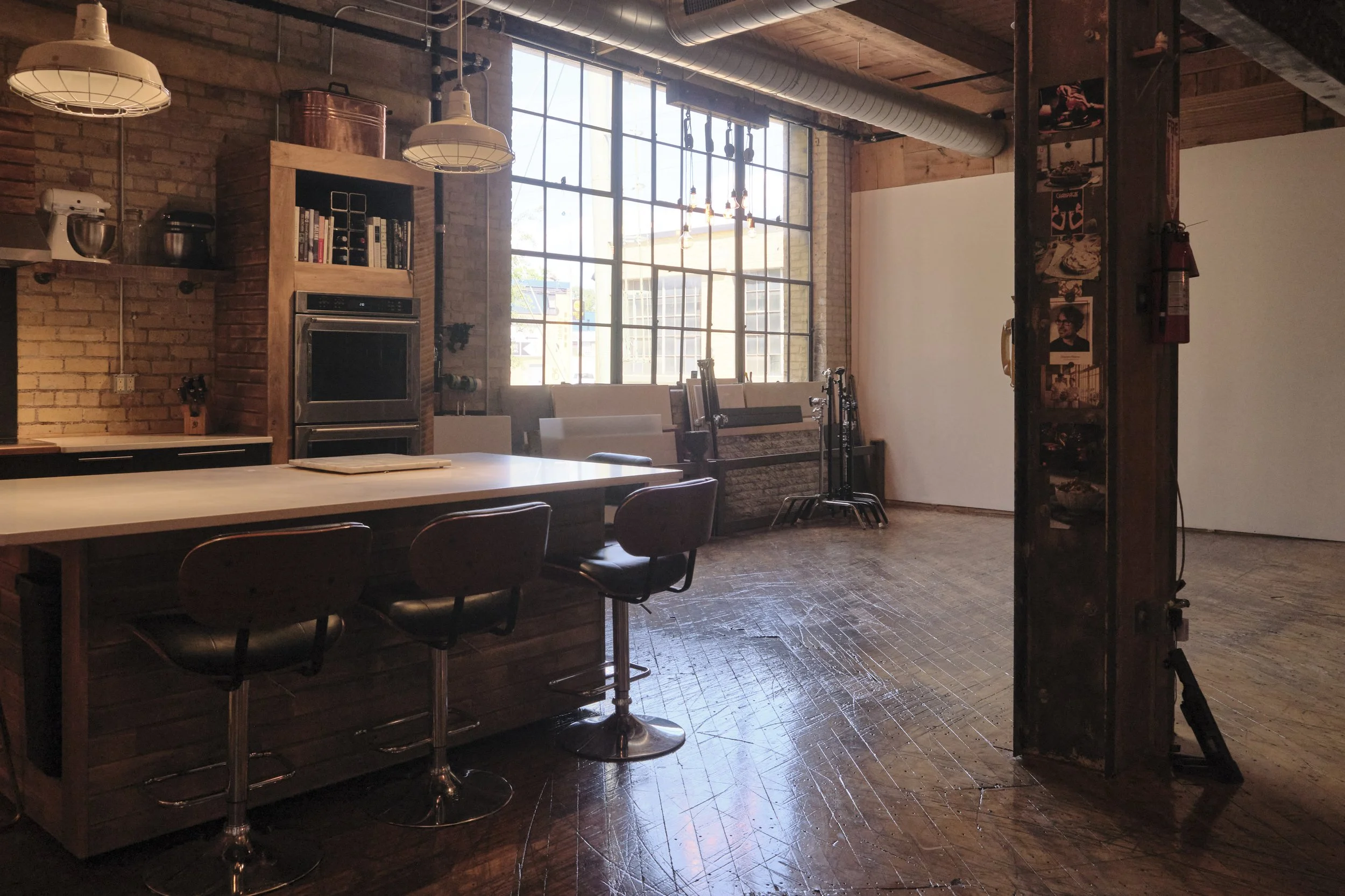 Interior of a spacious industrial-style kitchen and dining area with brick walls, large windows, a wooden table with chairs, and various kitchen appliances and equipment.