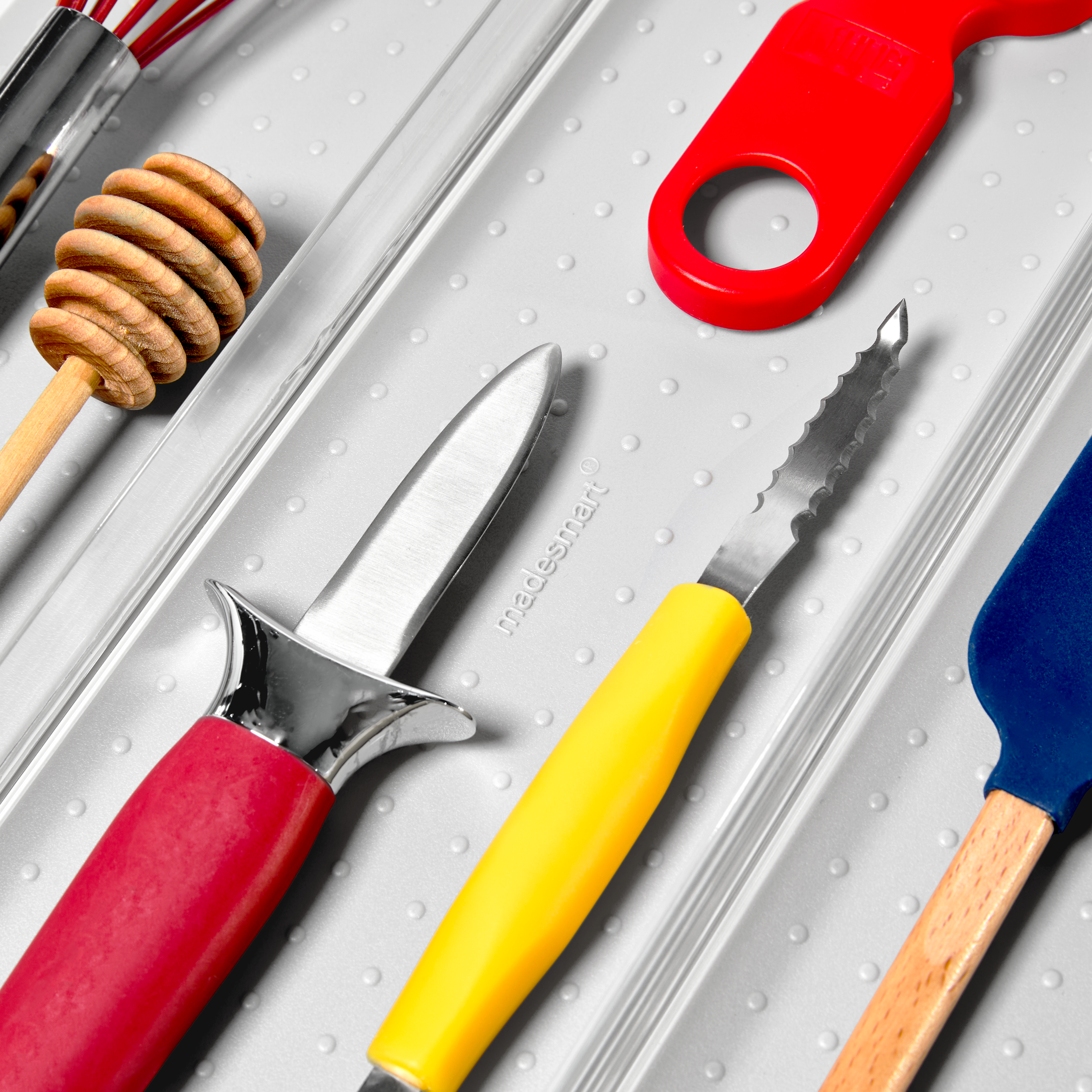 Collection of kitchen tools on a gray textured surface, including a honey dipper, a paring knife, a small serrated knife with a yellow handle, a red bottle opener, and a wooden-handled spatula with a blue cover.