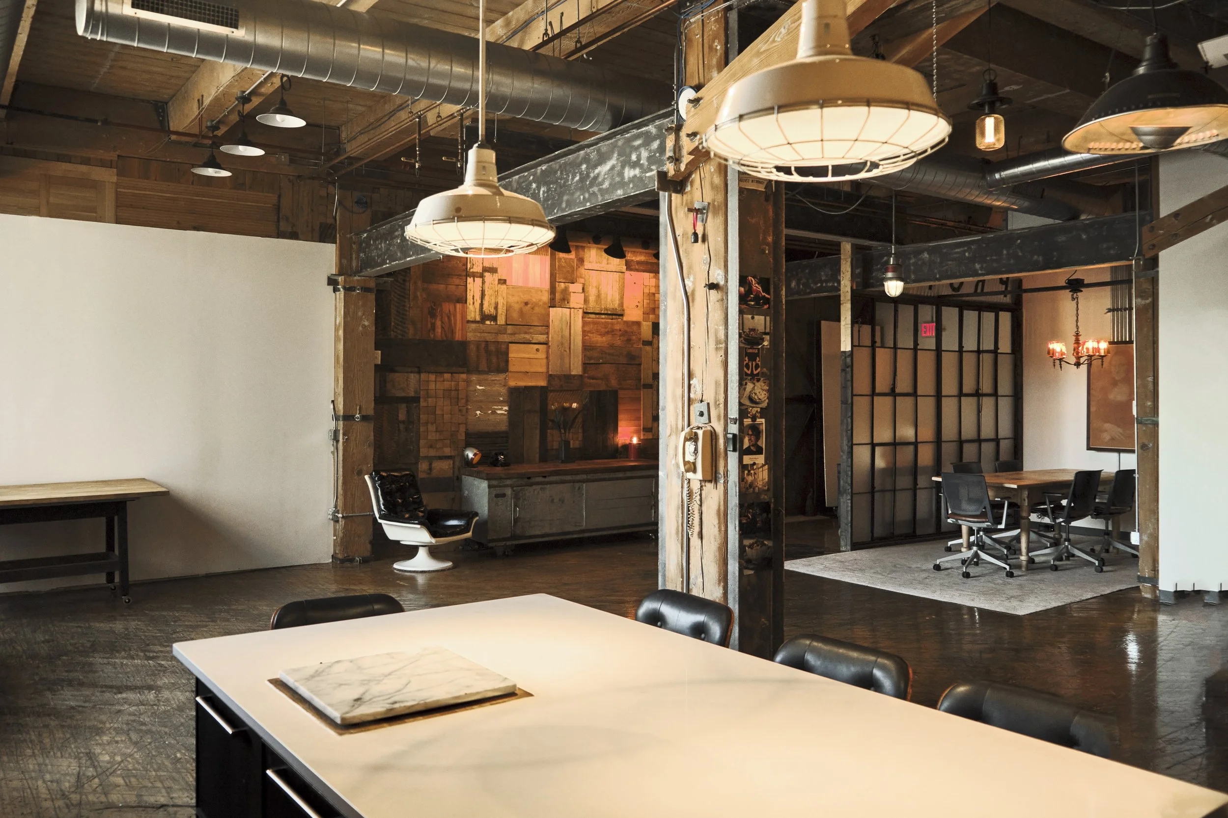 Interior of a modern office or coworking space with wooden and industrial design elements, including a large white table with black chairs, pendant lighting, and additional seating areas.