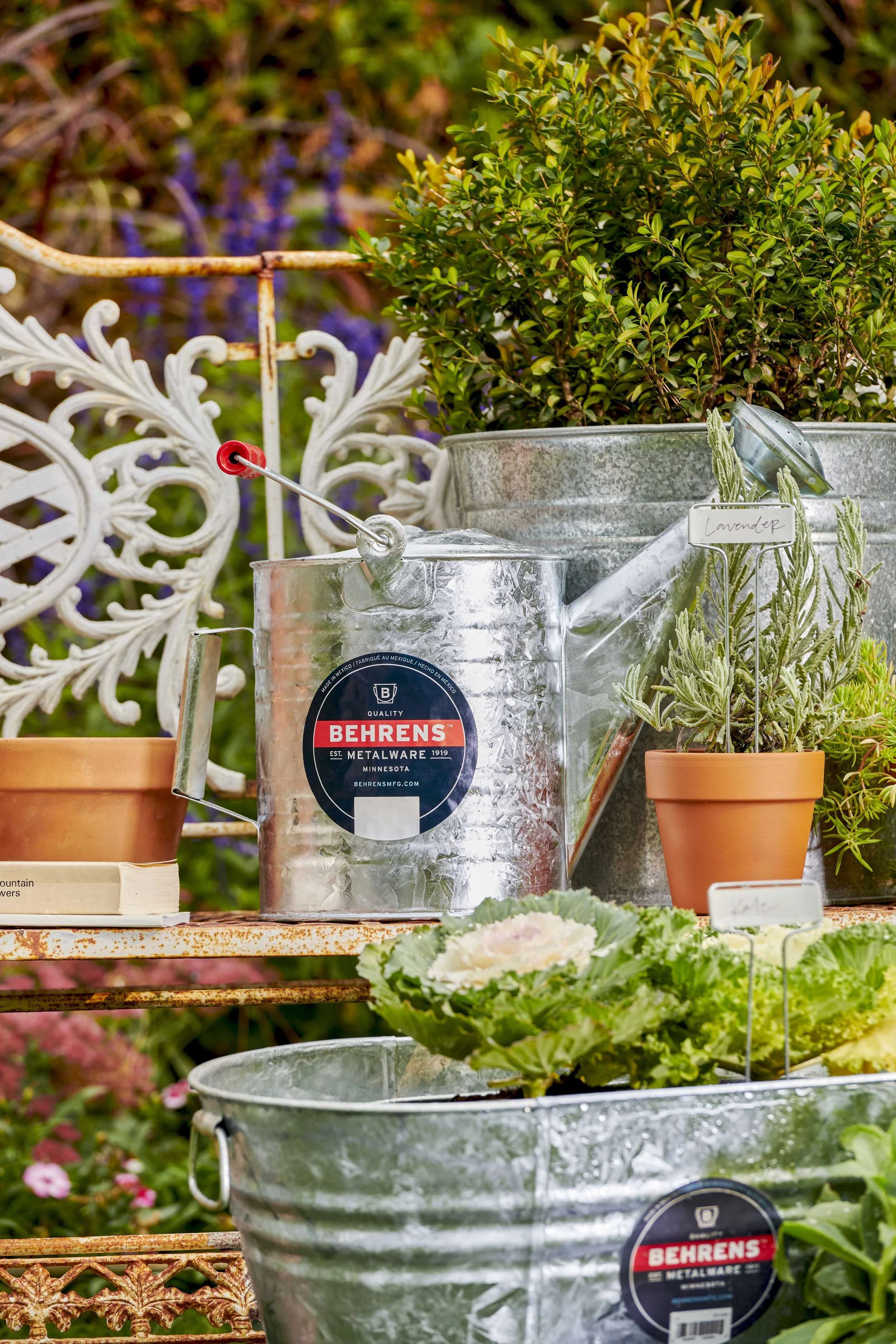 Various gardening supplies on a rusted metal shelf, including metal watering cans, potted lavender and other plants, and a garden book, with a decorative white metal chair back and greenery in the background.
