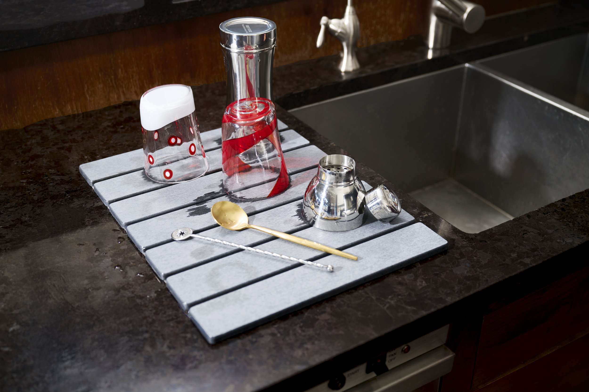 A kitchen countertop with a drying mat holding various glassware, a gold and silver spoon, and a cocktail shaker, next to a sink.