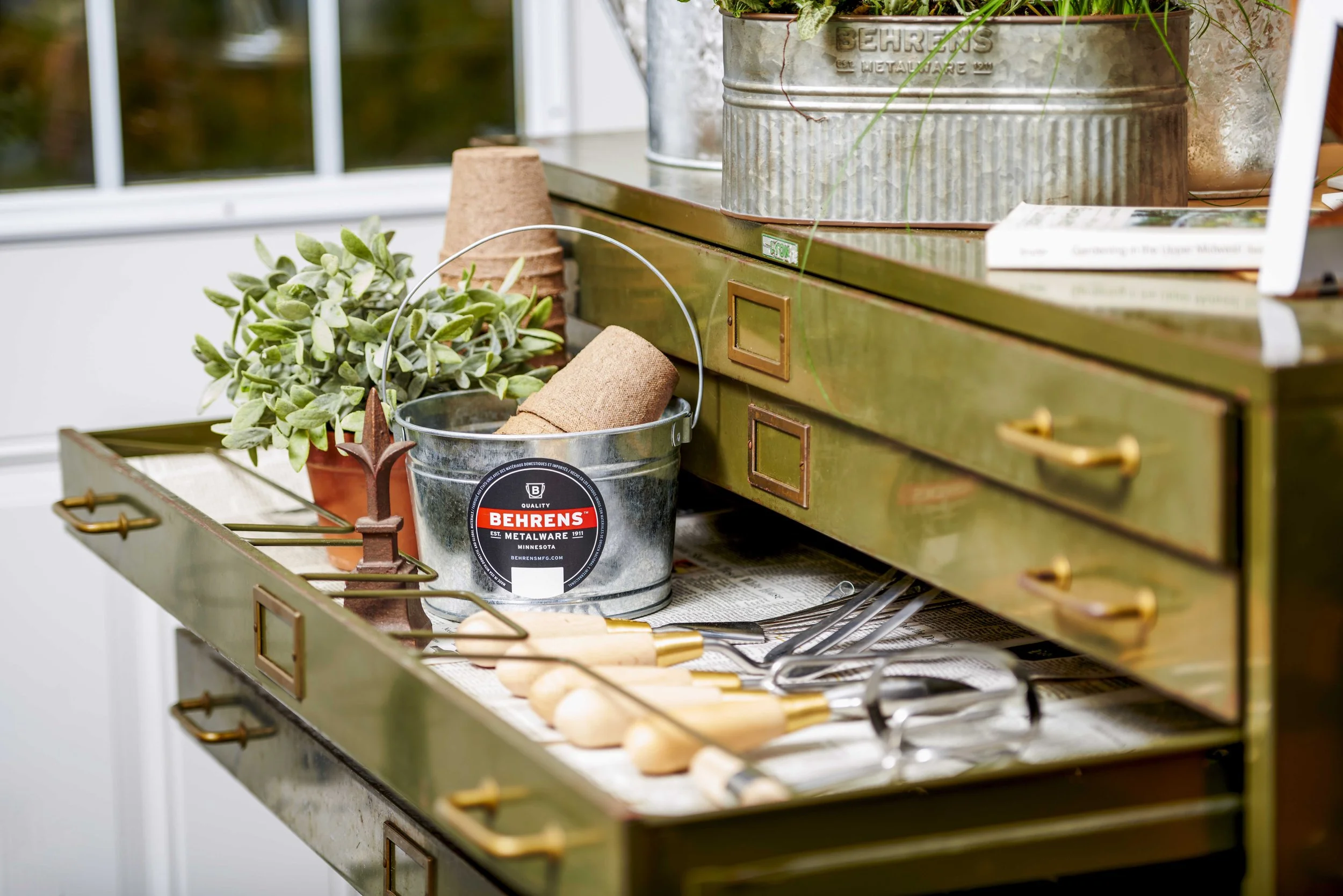 A vintage green metal cabinet with drawers, open to reveal gardening tools with wooden handles, a potted plant, a small metal bucket with a roll of twine, a pair of black glasses, and a newspaper underneath the tools.