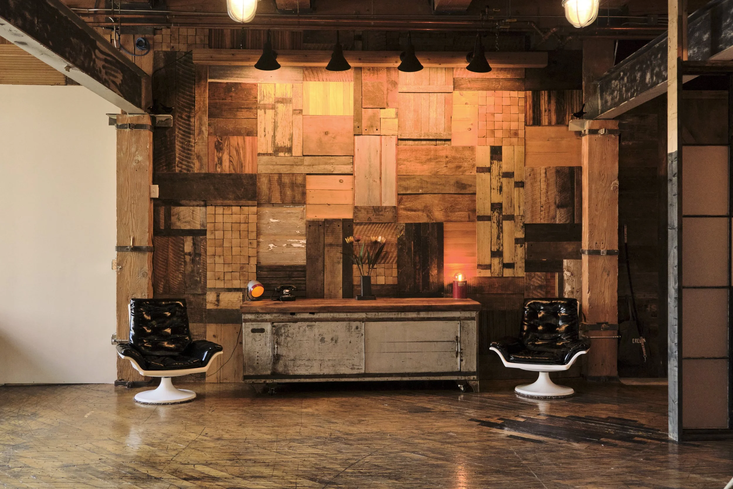 Interior of a room with a wooden patchwork wall, vintage desk, two black leather chairs, a candle, and a flower arrangement.