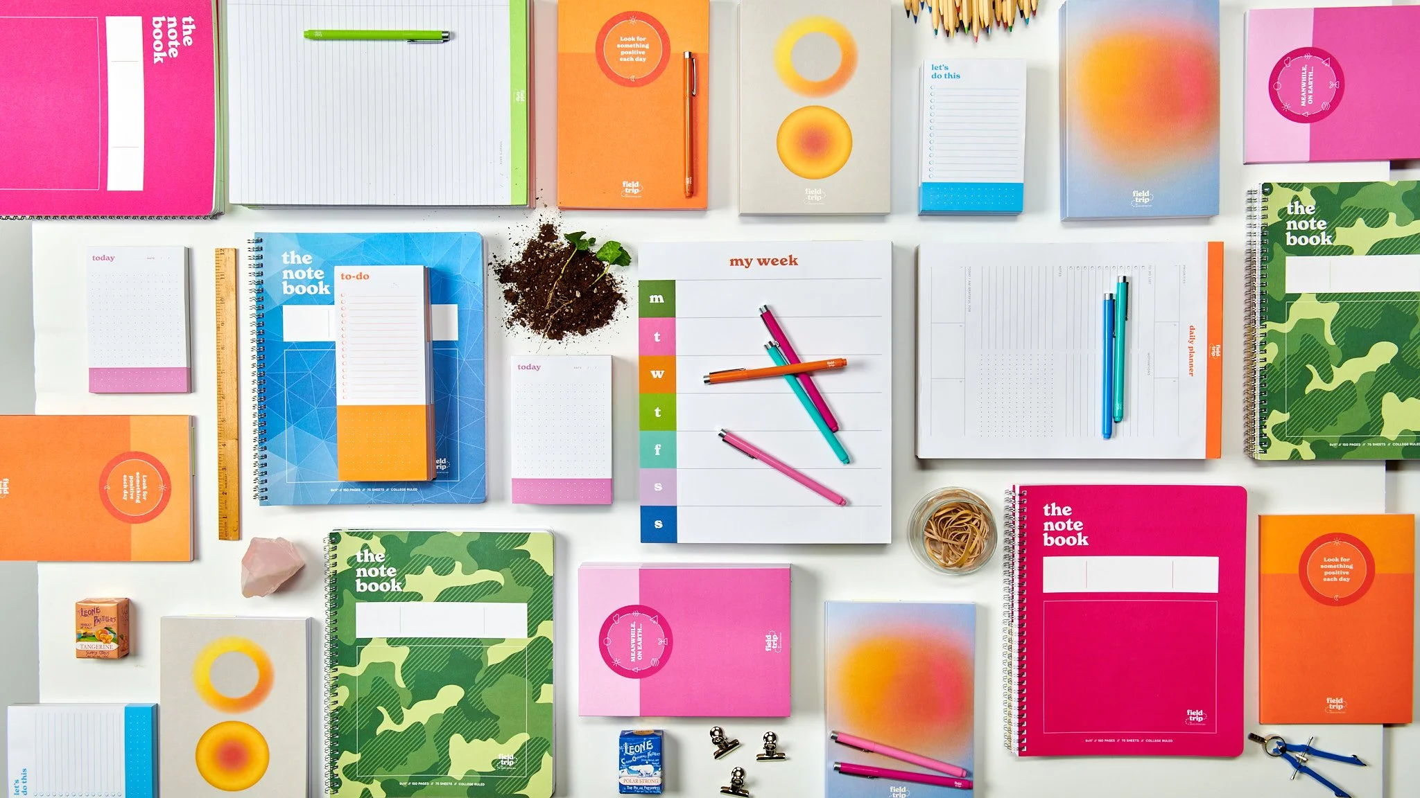 Spread of various colorful notebooks, planners, pens, and stationery items arranged on a white surface, including notebooks in pink, orange, blue, green, and patterned covers, along with markers, clips, a ruler, a small pile of soil, and a pink cryst