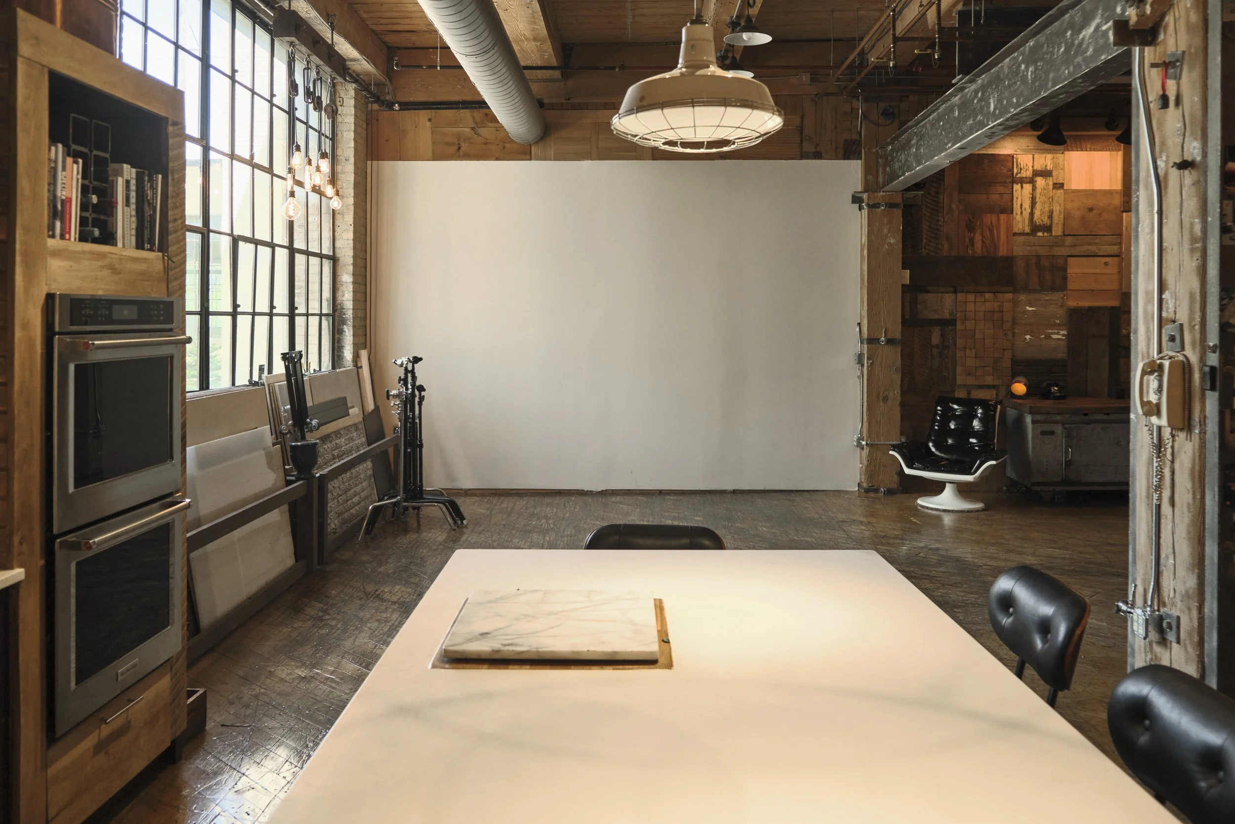 Interior of a rustic-style photography studio or office with a large white backdrop, wooden walls and ceiling, a large window with multiple panes, a table with a marble panel, black chairs, studio equipment, and a black modern lounge chair.
