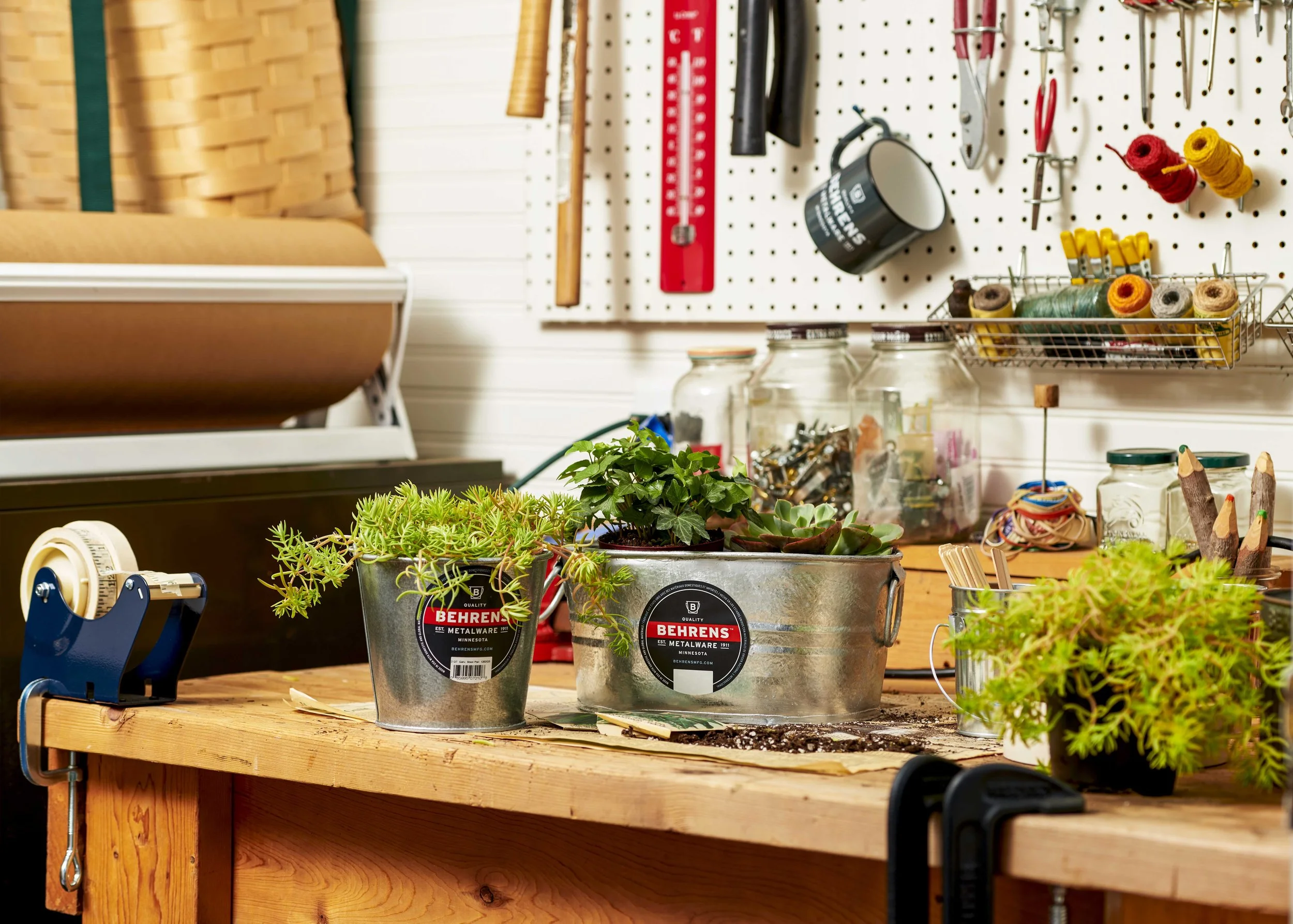A workshop table with potted succulents, tools, and supplies for gardening and crafting. A pegboard with tools, jars, and rolled yarns is in the background.