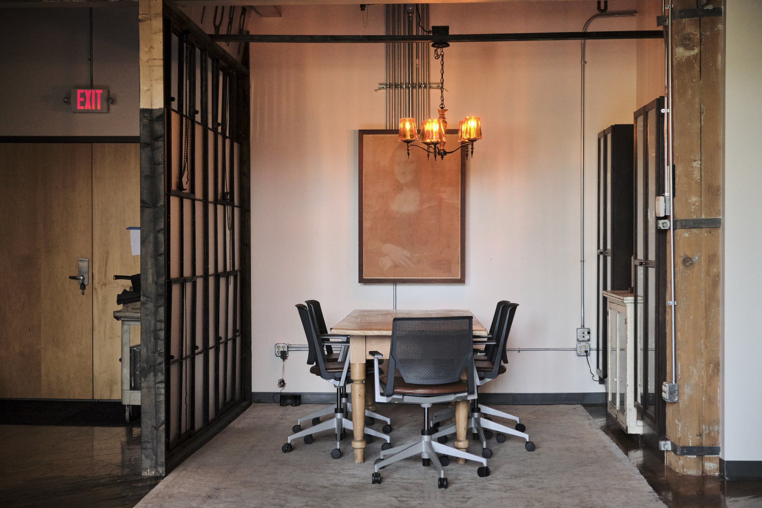 A modern office conference room with a wooden table surrounded by five office chairs, a chandelier with five orange lamps, a framed cork bulletin board, and a mix of industrial and rustic design elements. There is a door on the left and some visible 