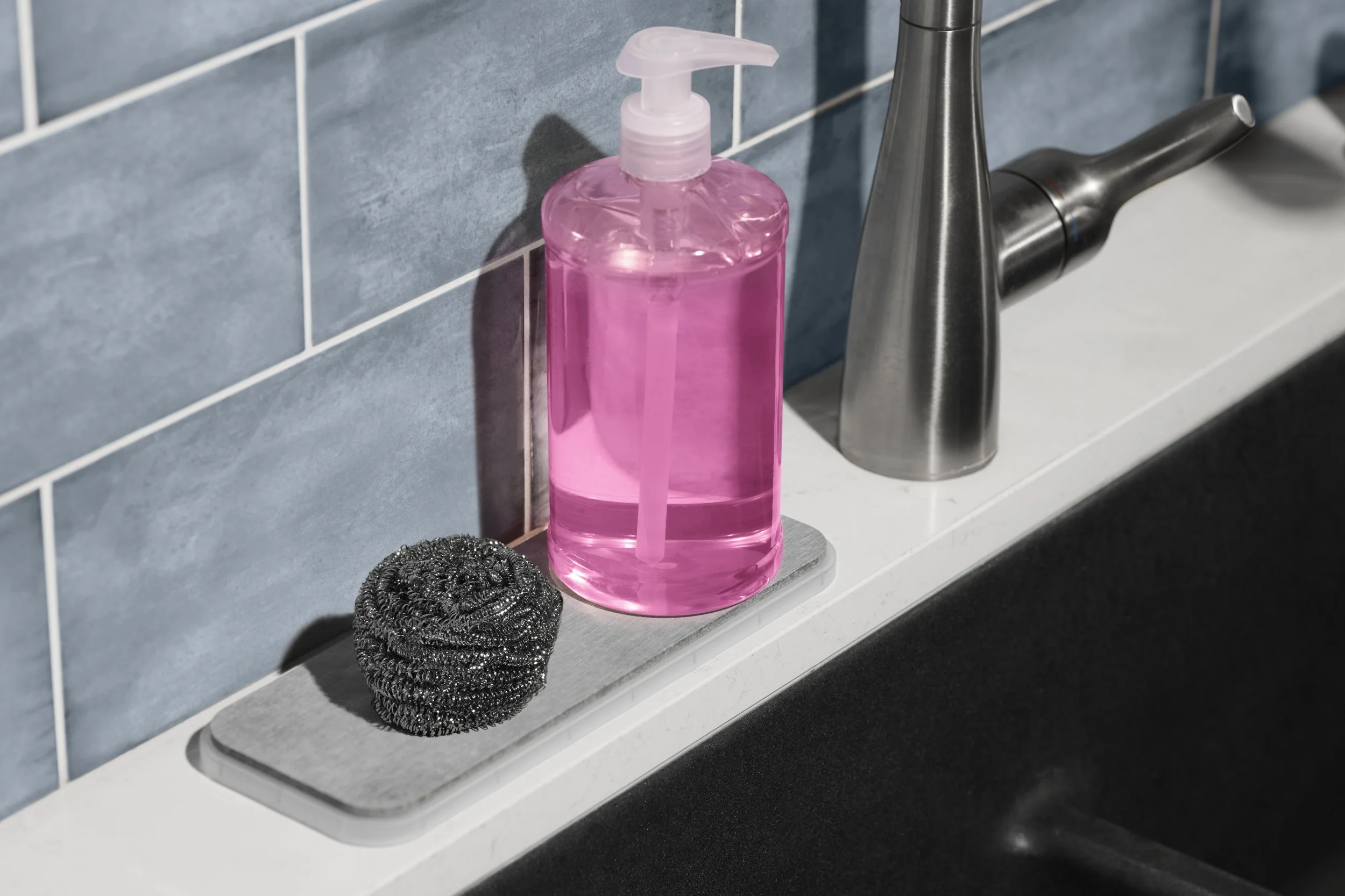A pink liquid soap dispenser, a black scrubbing sponge, and a silver kitchen faucet on a white countertop.