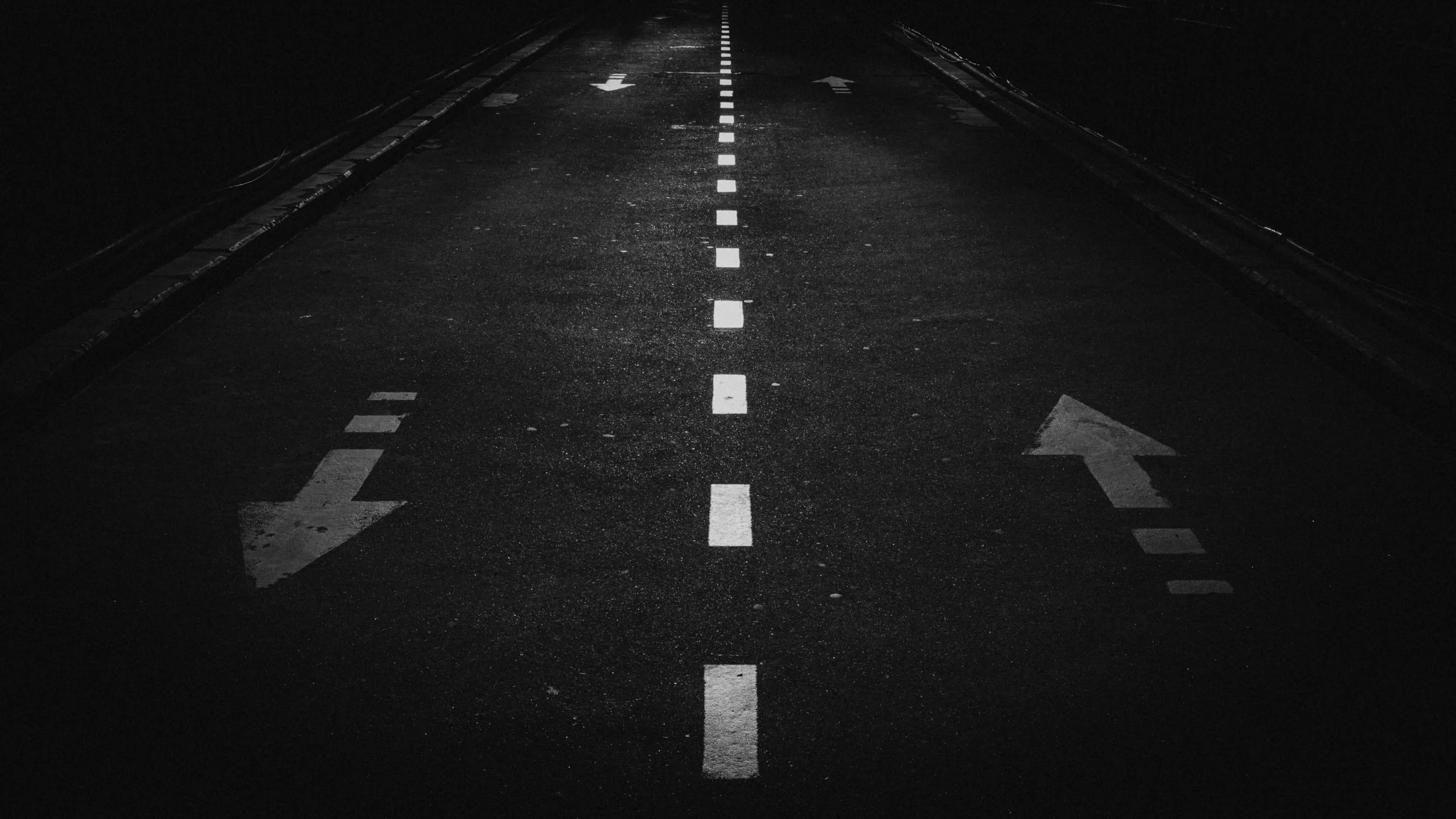 Dark road at night with white dashed center line and arrows indicating directions, one arrow pointing left and the other pointing right.
