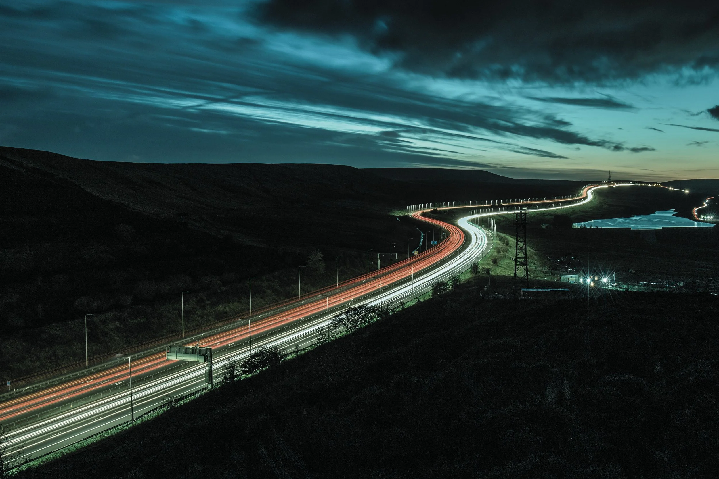 Long exposure photograph of a winding highway at night with light trails from vehicles, hills in the background, and a dark sky with clouds.