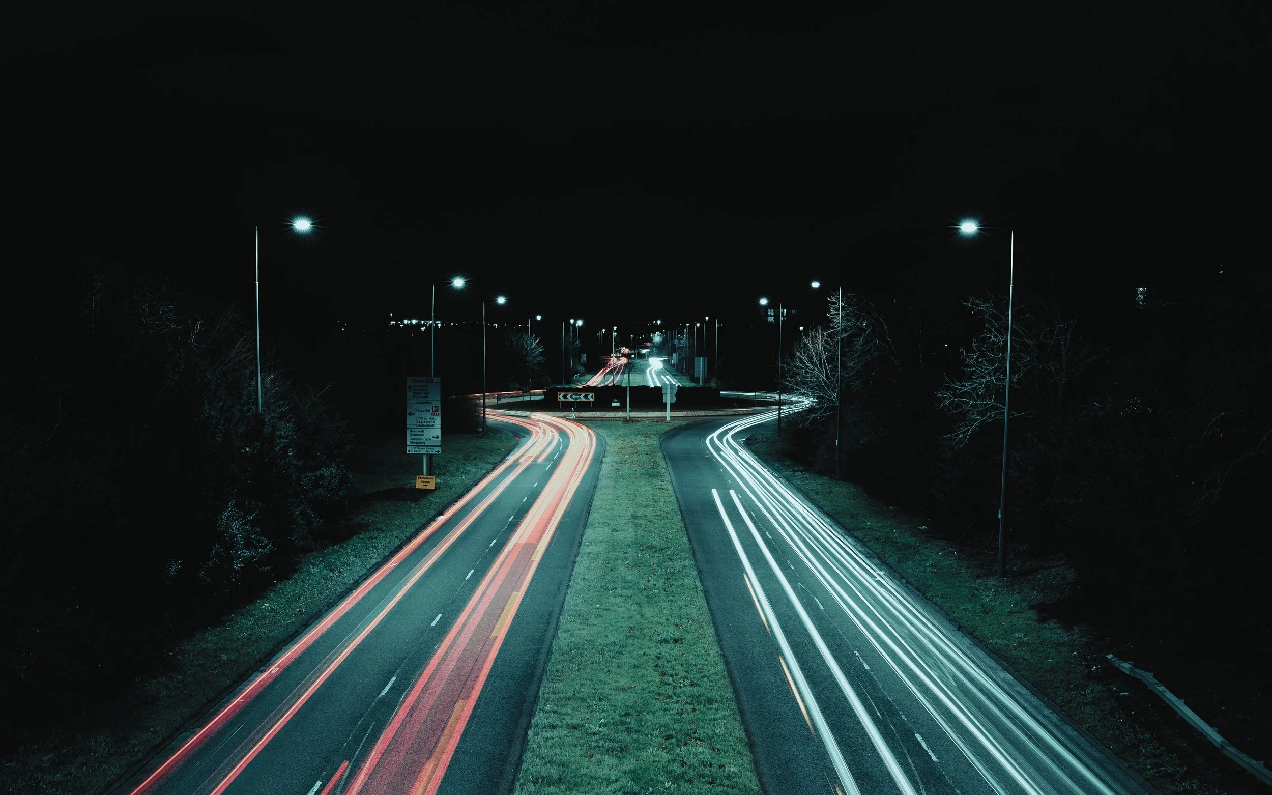 Night scene of a two-lane road with light trails from passing vehicles, streetlights illuminating the scene, and trees on both sides, taken with long exposure.