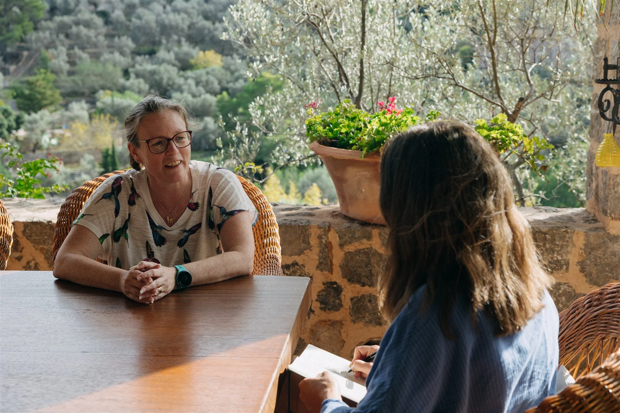 Two women having a conversation at a wooden table on a terrace with a scenic view of greenery and trees in the background.