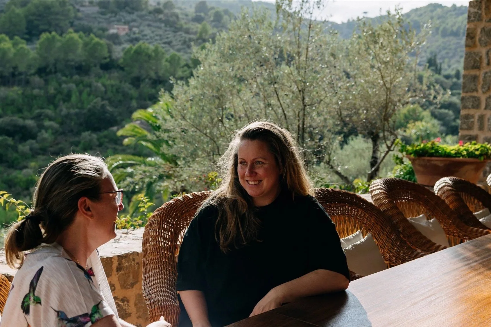 Two women sitting at a wooden table outdoors, facing each other and smiling, with a scenic green hillside and trees in the background.