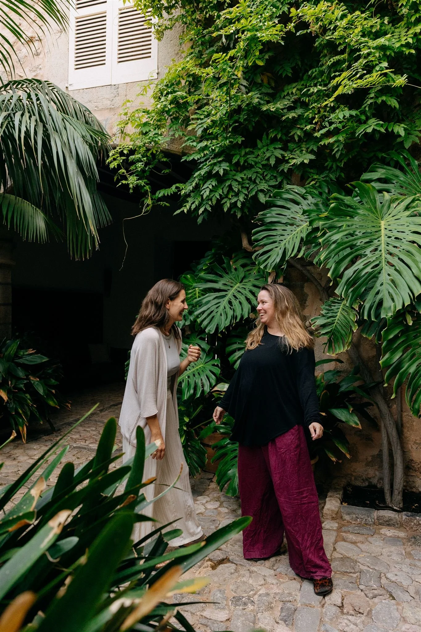 Two women smiling and talking in a lush, green garden with large tropical leaves and stone pavement.