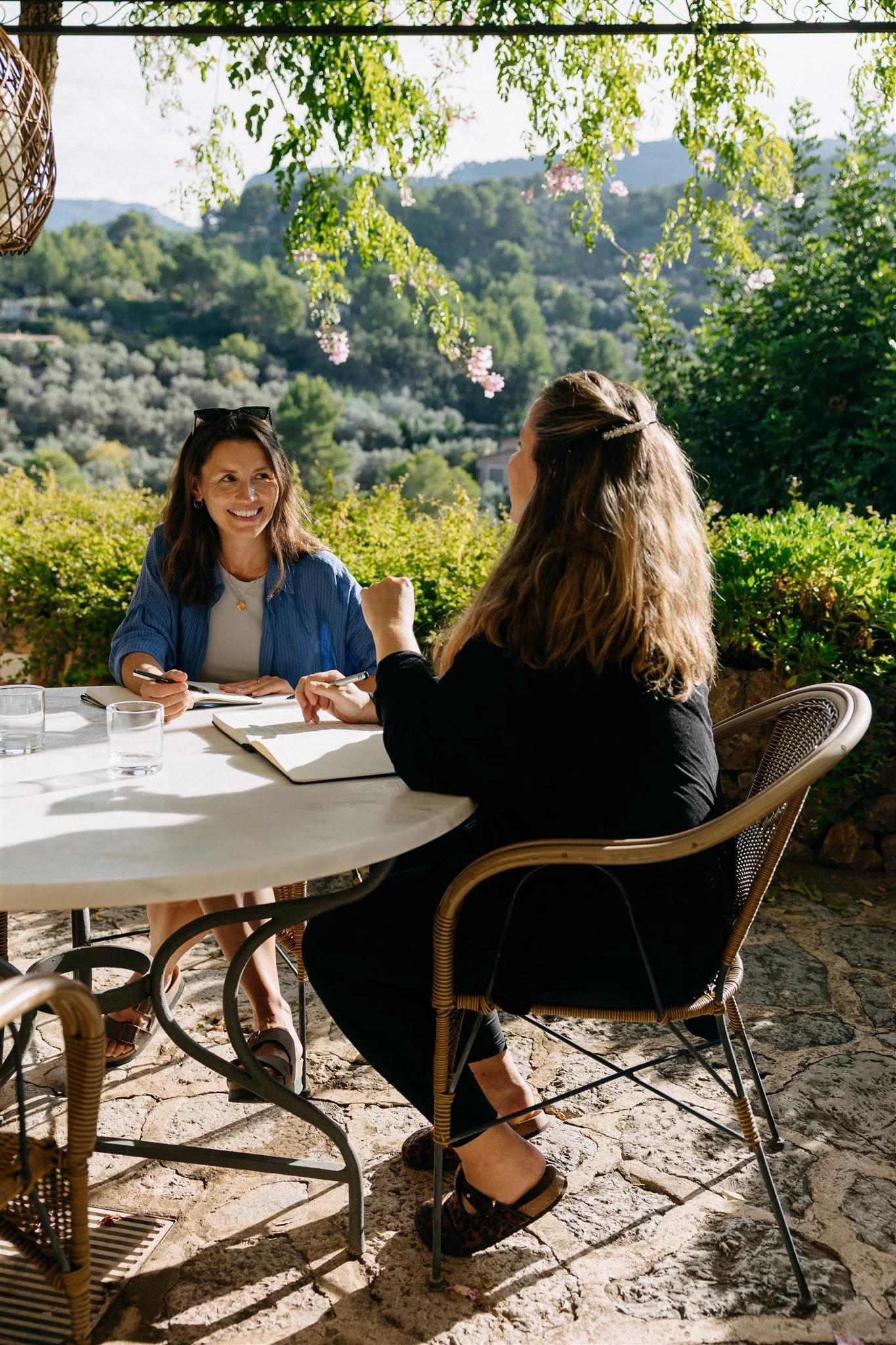 Two women having a conversation outdoors at a round table, surrounded by lush greenery and mountains in the background, with sunlight filtering through trees.