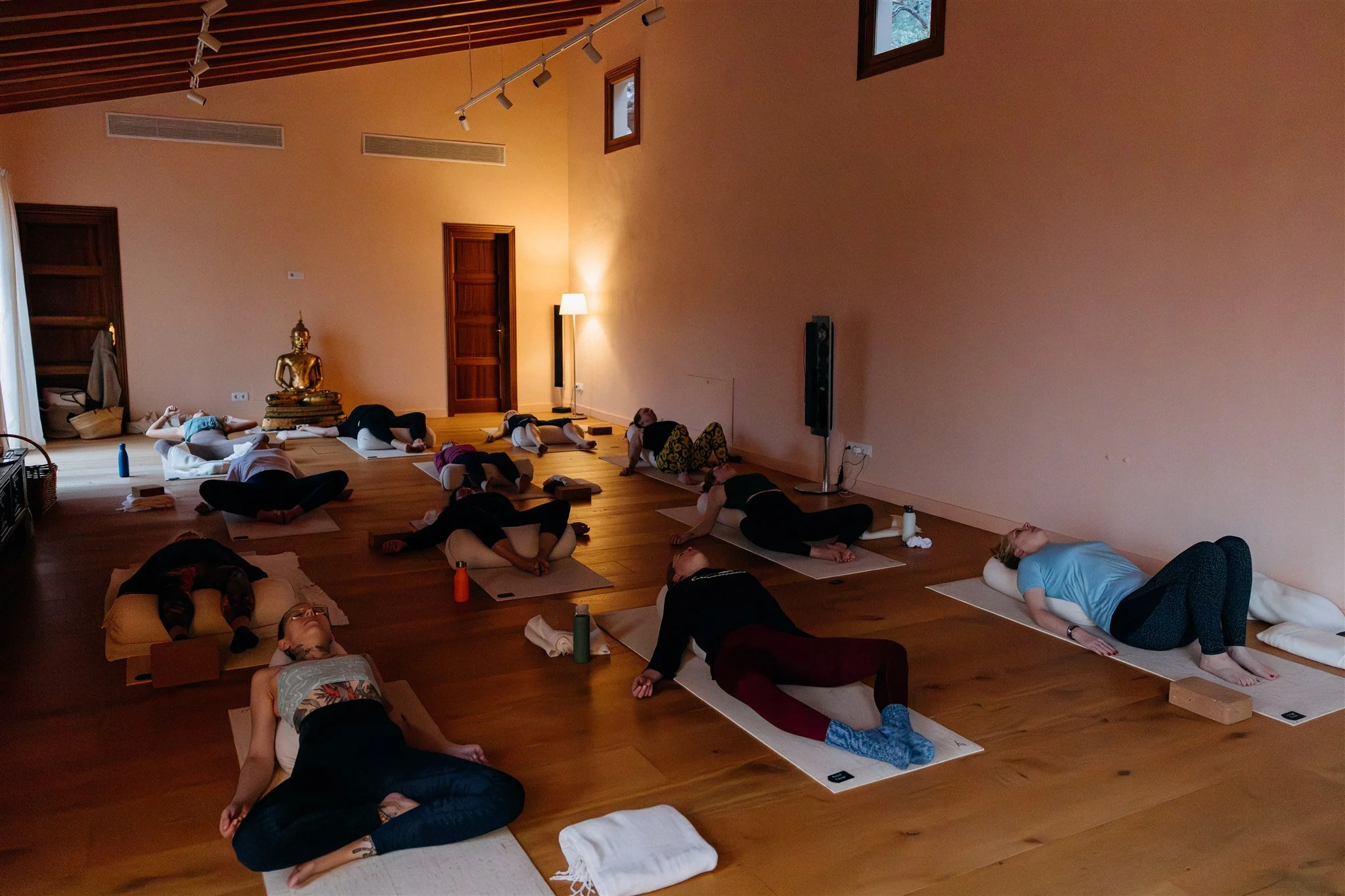 People participating in a yoga class, lying on their mats on a wooden floor, in a dimly lit room with soft lighting and a Buddha statue.
