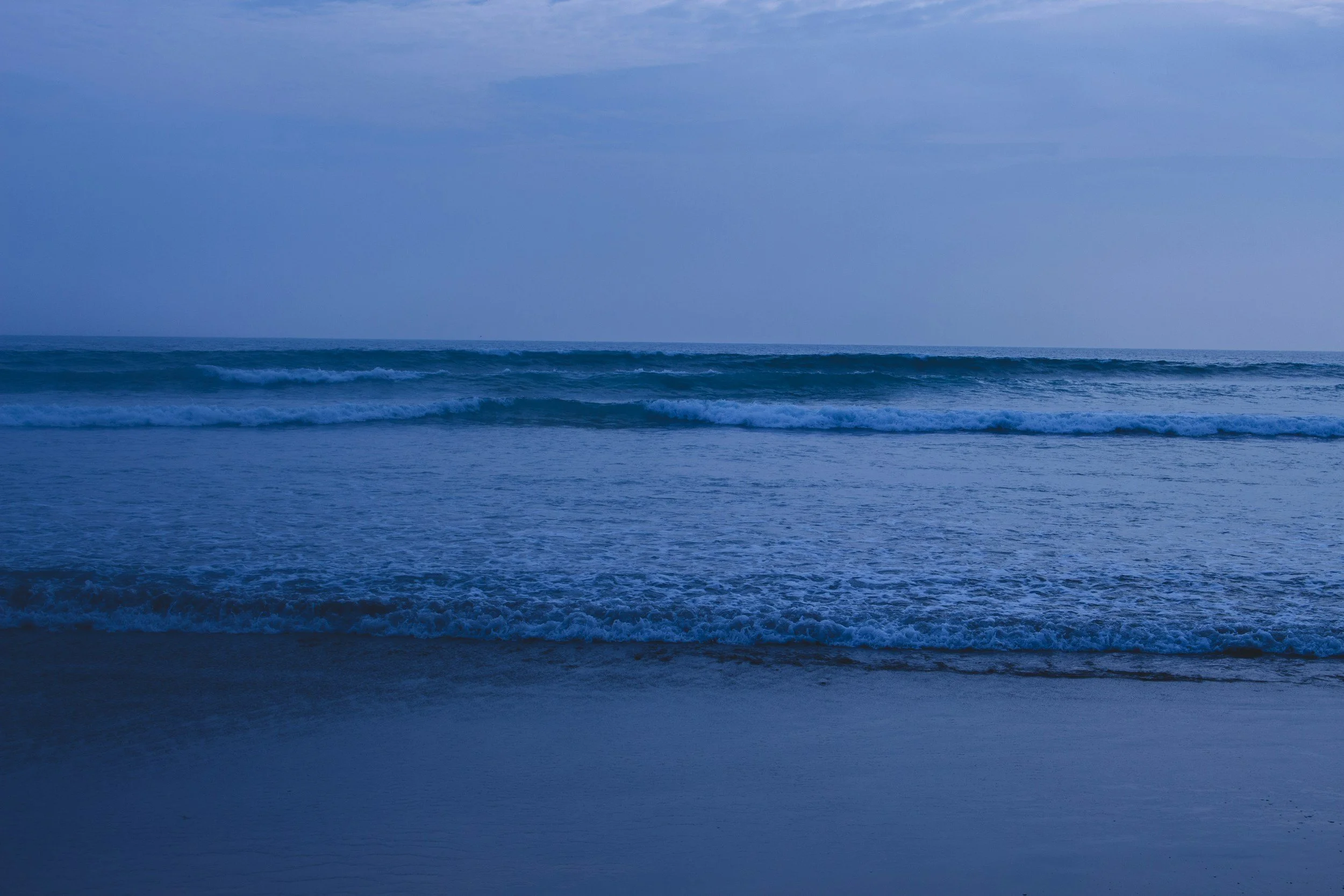 Calm ocean waves breaking on the shore at dusk under a cloudy sky.