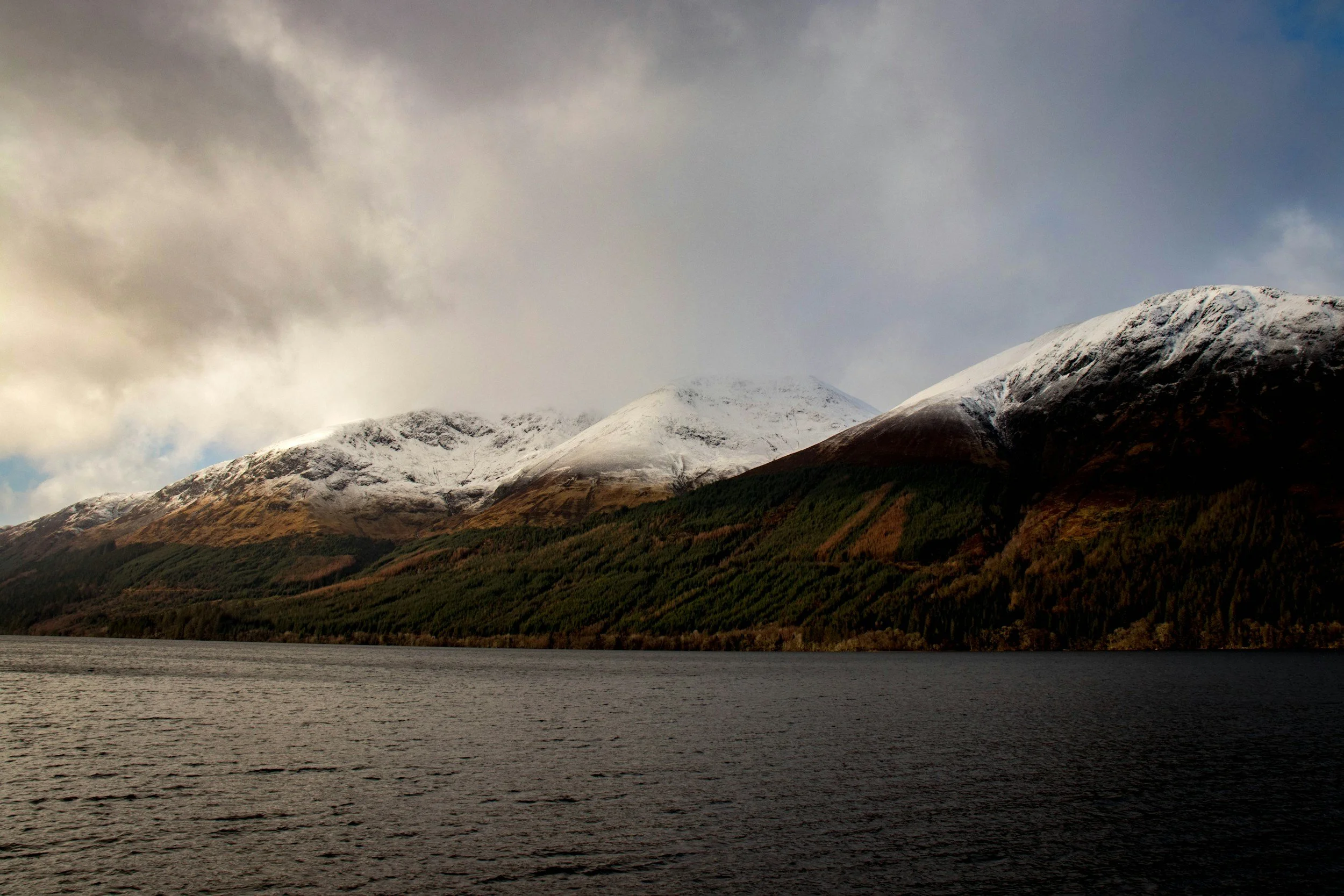 Snow-capped mountains with green and brown slopes and a dark lake in the foreground, under a cloudy sky.