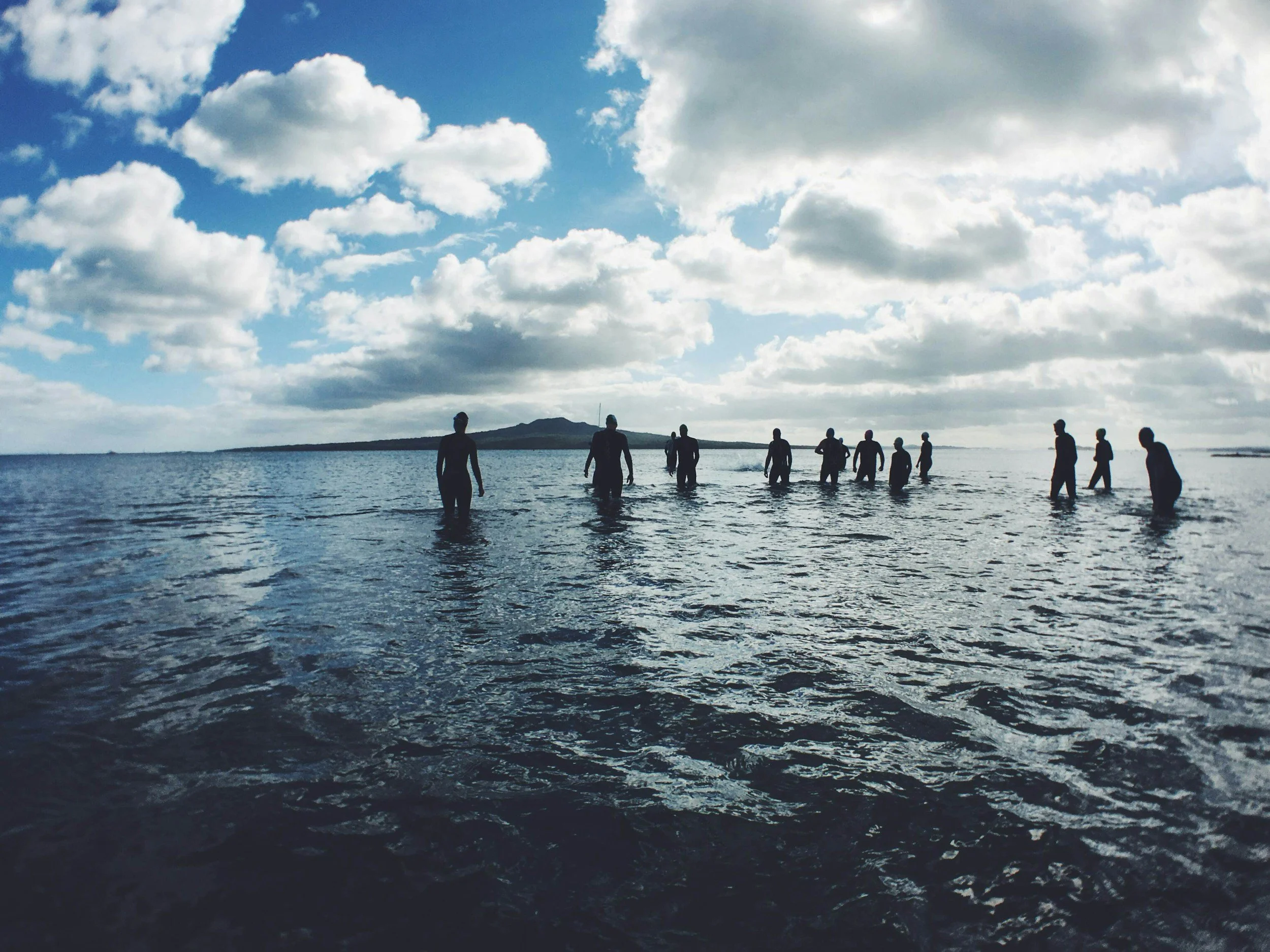 People wading in the ocean with a cloudy sky and distant land in the background.