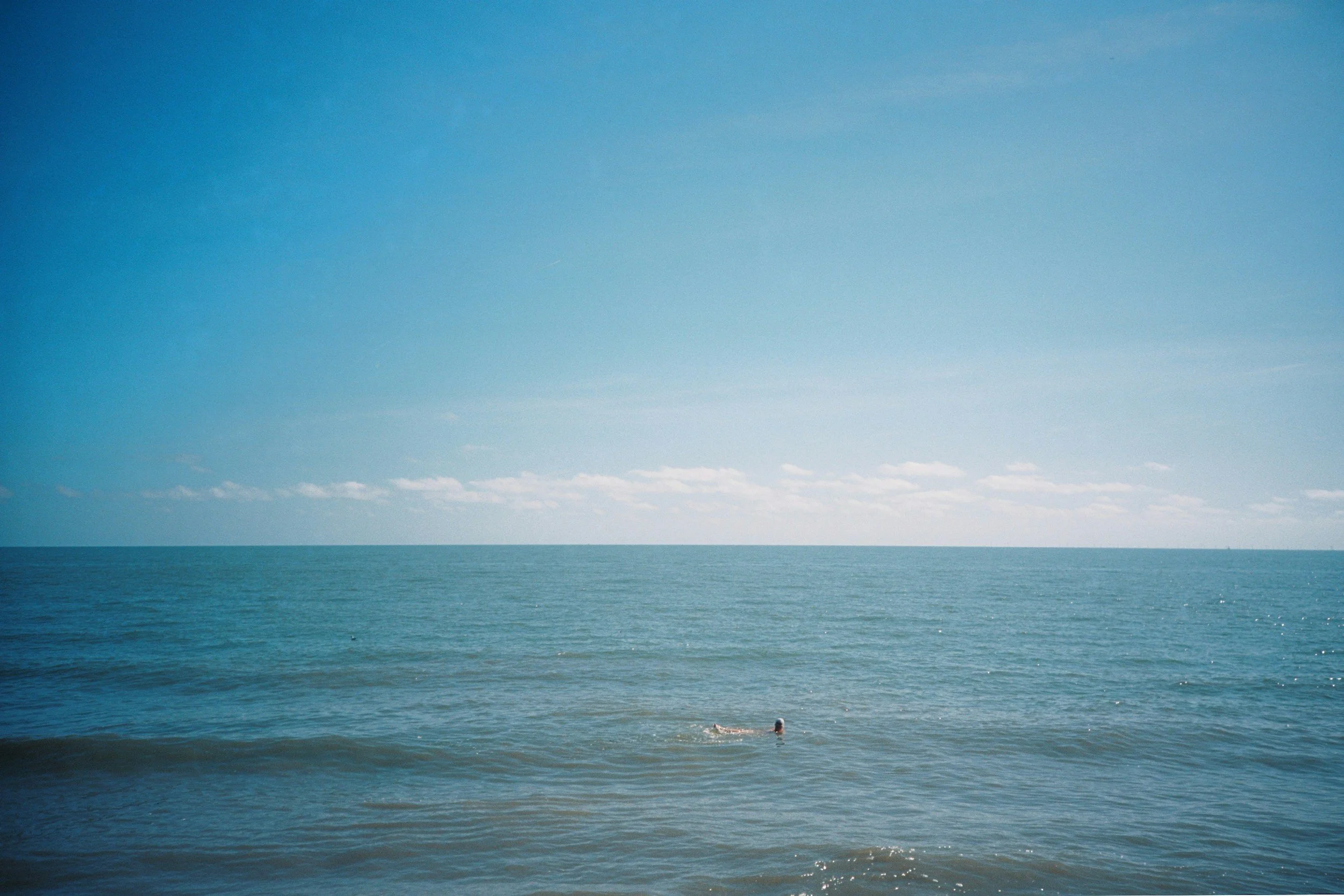 A person swimming in the ocean near the shore under a clear blue sky with some thin clouds.