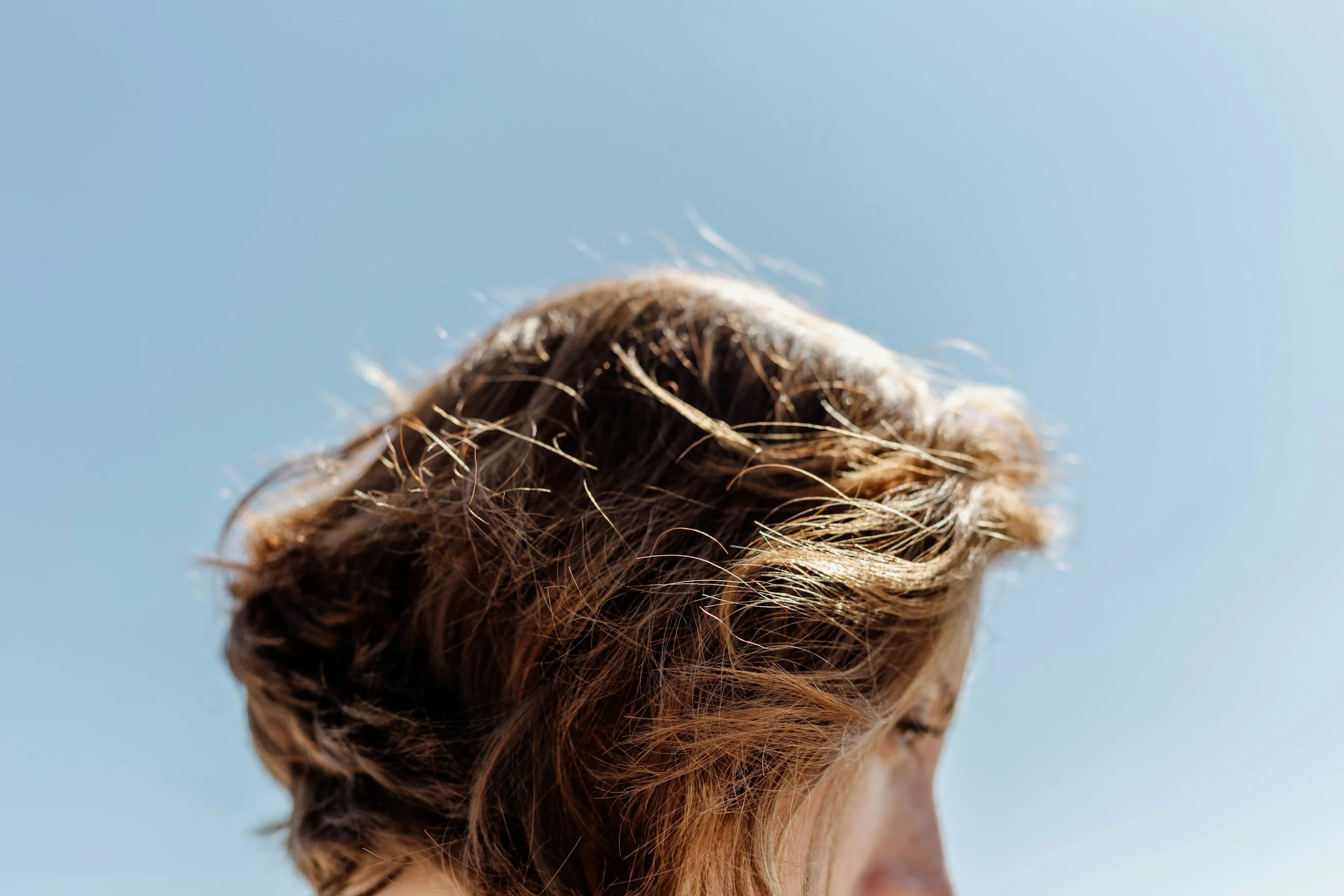 Close-up of a person's head with messy light brown hair against a clear blue sky.