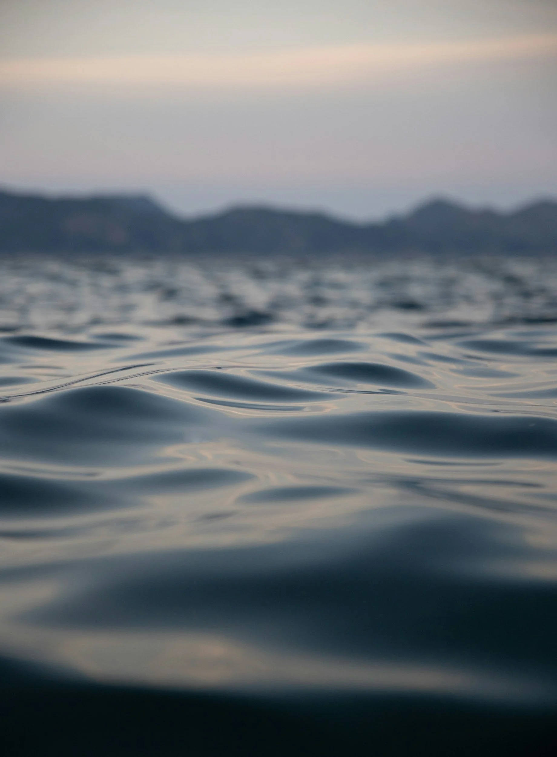Close-up of calm ocean water with distant silhouette of mountains and a dusky sky.