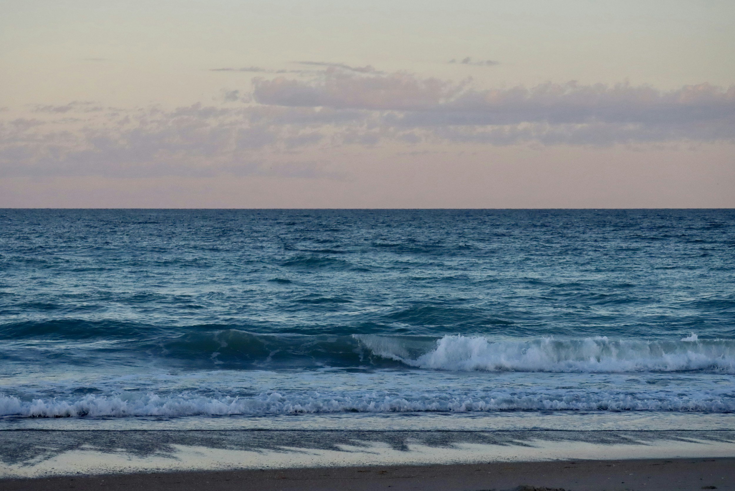 A peaceful view of the ocean with small waves approaching the sandy shore and a partly cloudy sky overhead.
