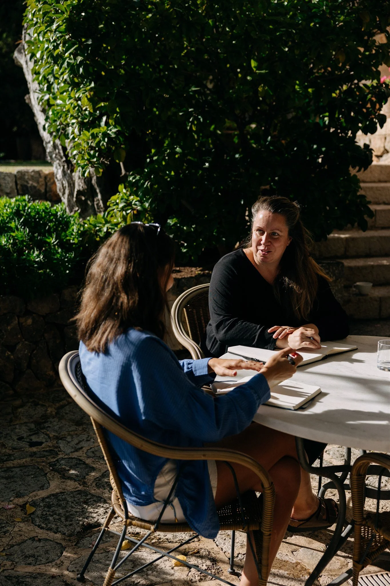 Two women sitting at an outdoor table having a conversation, surrounded by greenery