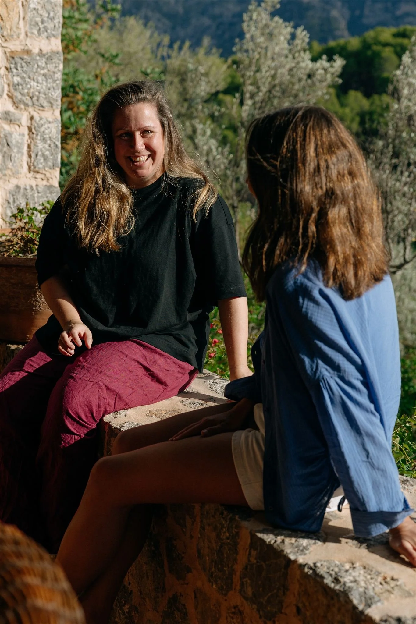 Two women sitting outside on a stone ledge, engaging in conversation with lush green trees and mountains in the background.