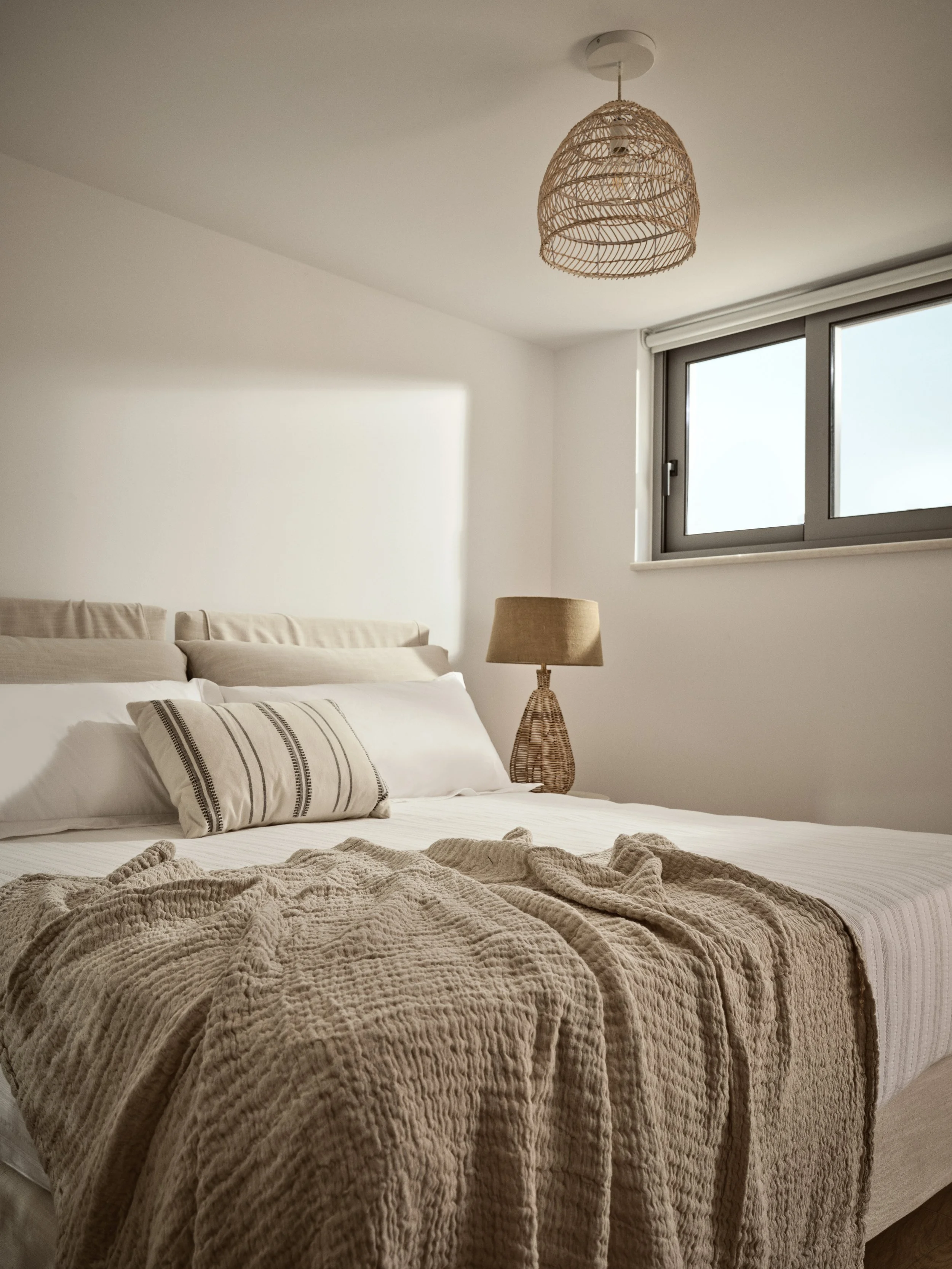A cozy bedroom with a neatly made bed, beige pillows with striped patterns, a textured beige blanket, a bedside table with a wicker base lamp, a window with a blind, and a rattan ceiling light fixture.