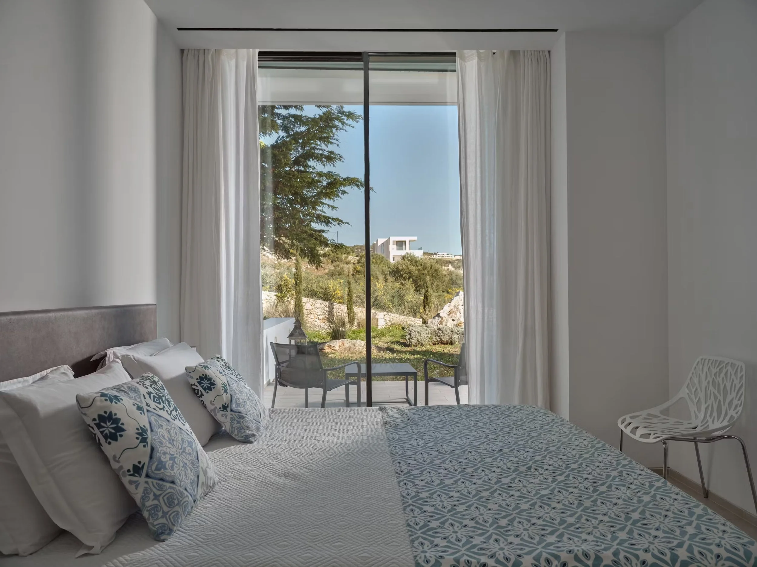 A bedroom with a large window showing trees and buildings outside, white curtains, a bed with white and patterned pillows, and a white chair.