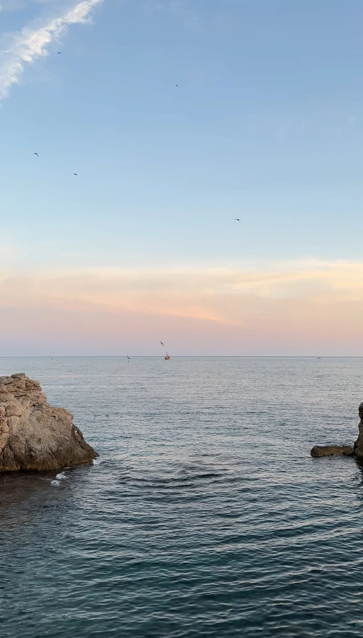 A serene ocean view at sunset with calm water, rocky formations on both sides, a few sailboats on the horizon, and a pastel-colored sky.