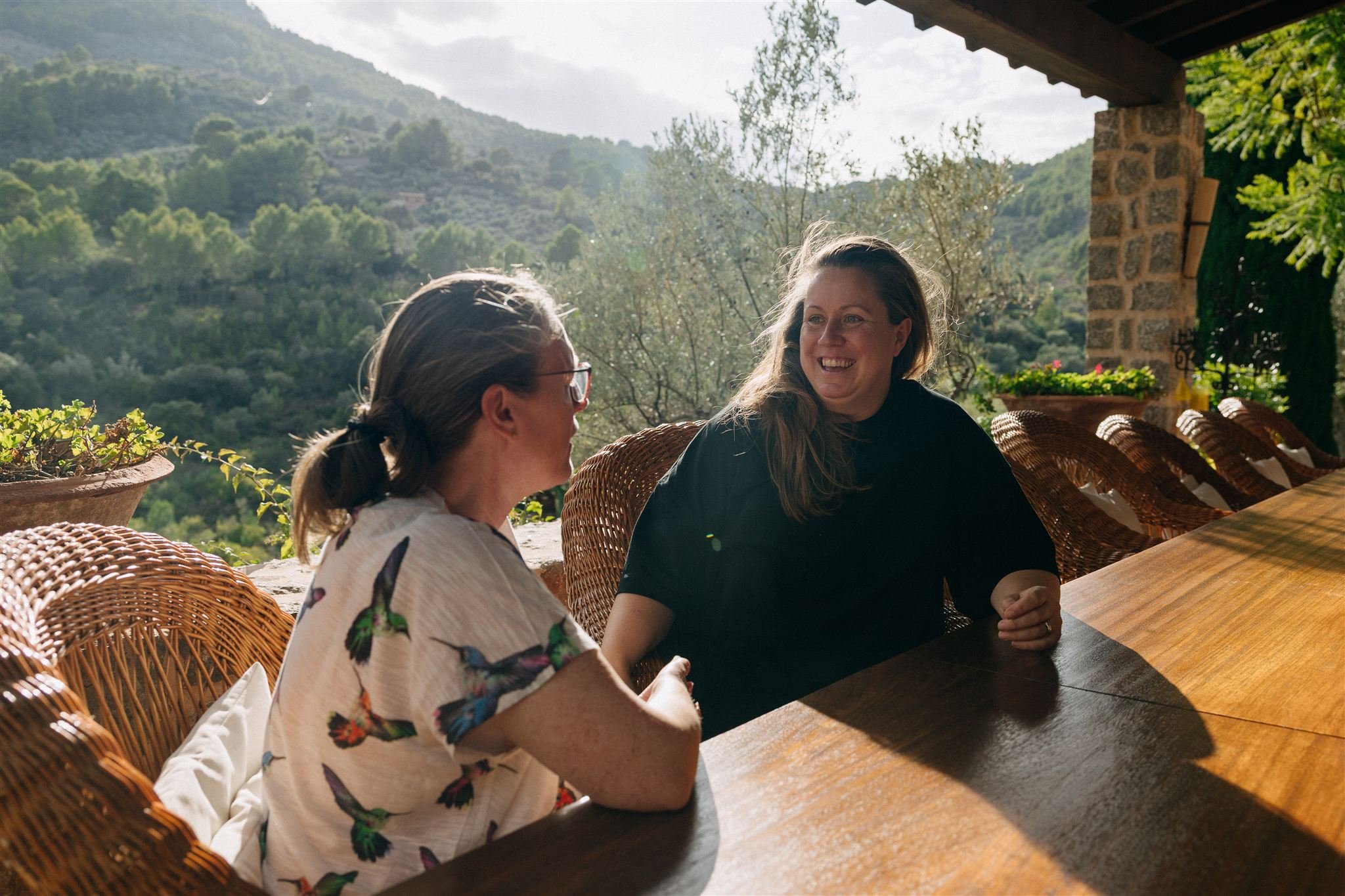 Two women sitting at a wooden outdoor table, smiling and talking, with a scenic mountain and green landscape background.