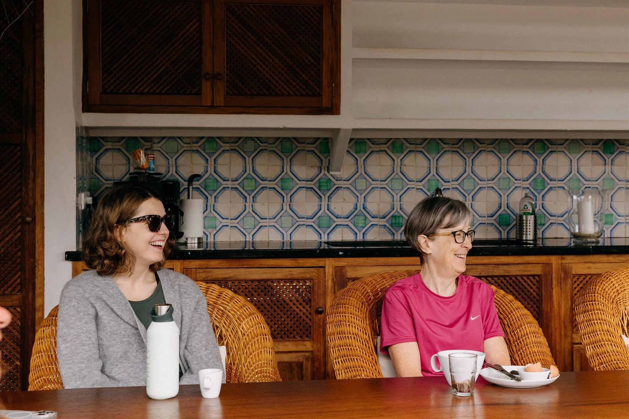 Two women sitting at a dining table, smiling and laughing. The woman on the left wears sunglasses, a gray sweater, and has a water bottle, while the woman on the right wears glasses and a red Nike T-shirt, with a plate of food and a glass of water in