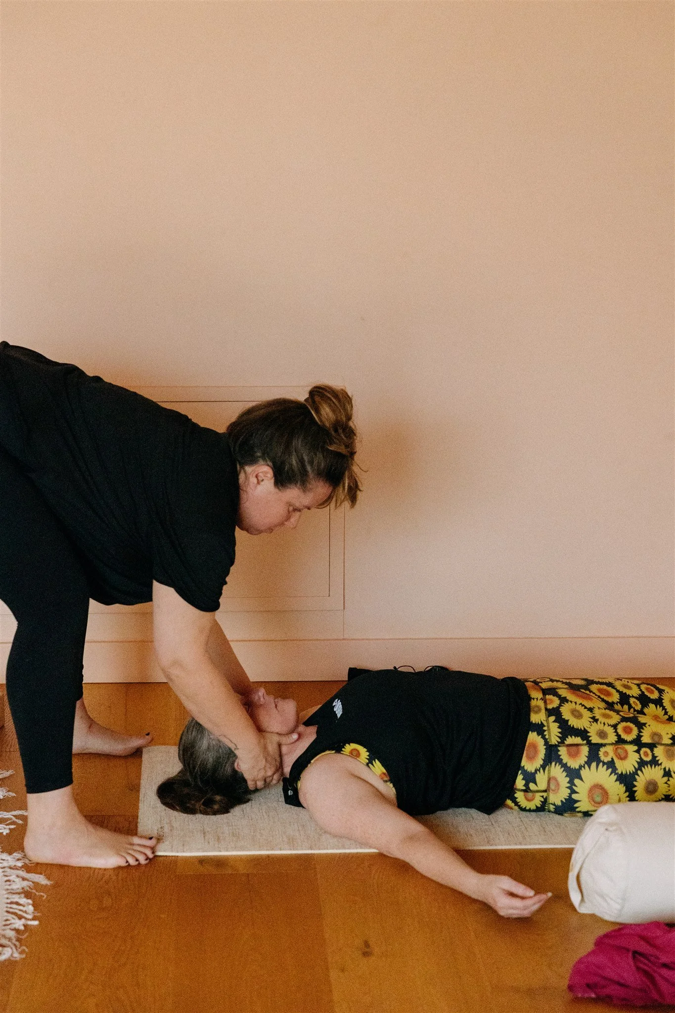 Person providing CPR to another person lying on the floor, with a woman in black and sunflower-patterned pants, in a room with wooden flooring and a beige wall.